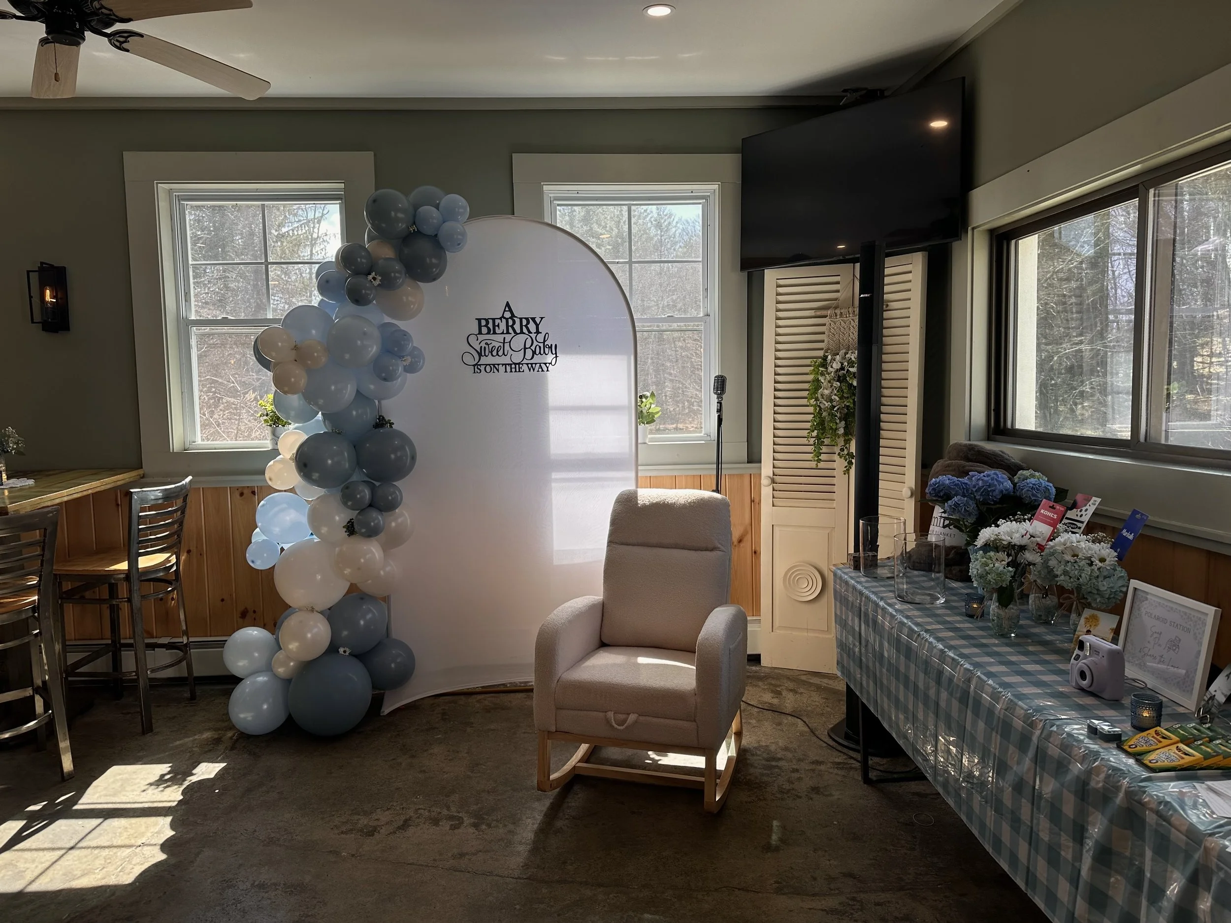 Interior of a room decorated for a baby shower with large balloon cluster and a white backdrop with text. There are two windows, a table with flowers and framed items, and various chairs.
