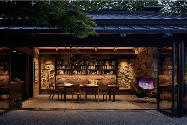 Interior view of a cozy dining area with a stone wall, bookshelf, guitar, and a fireplace, seen through large open glass doors.