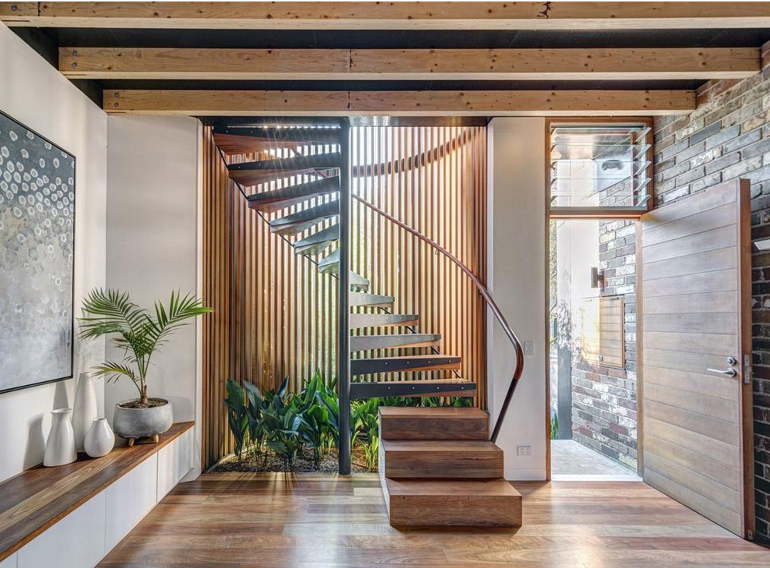 Interior view of a modern home featuring a wooden spiral staircase with a curved handrail, a large window, glass door, wooden floors, decorative potted plants, and minimalist wall art.