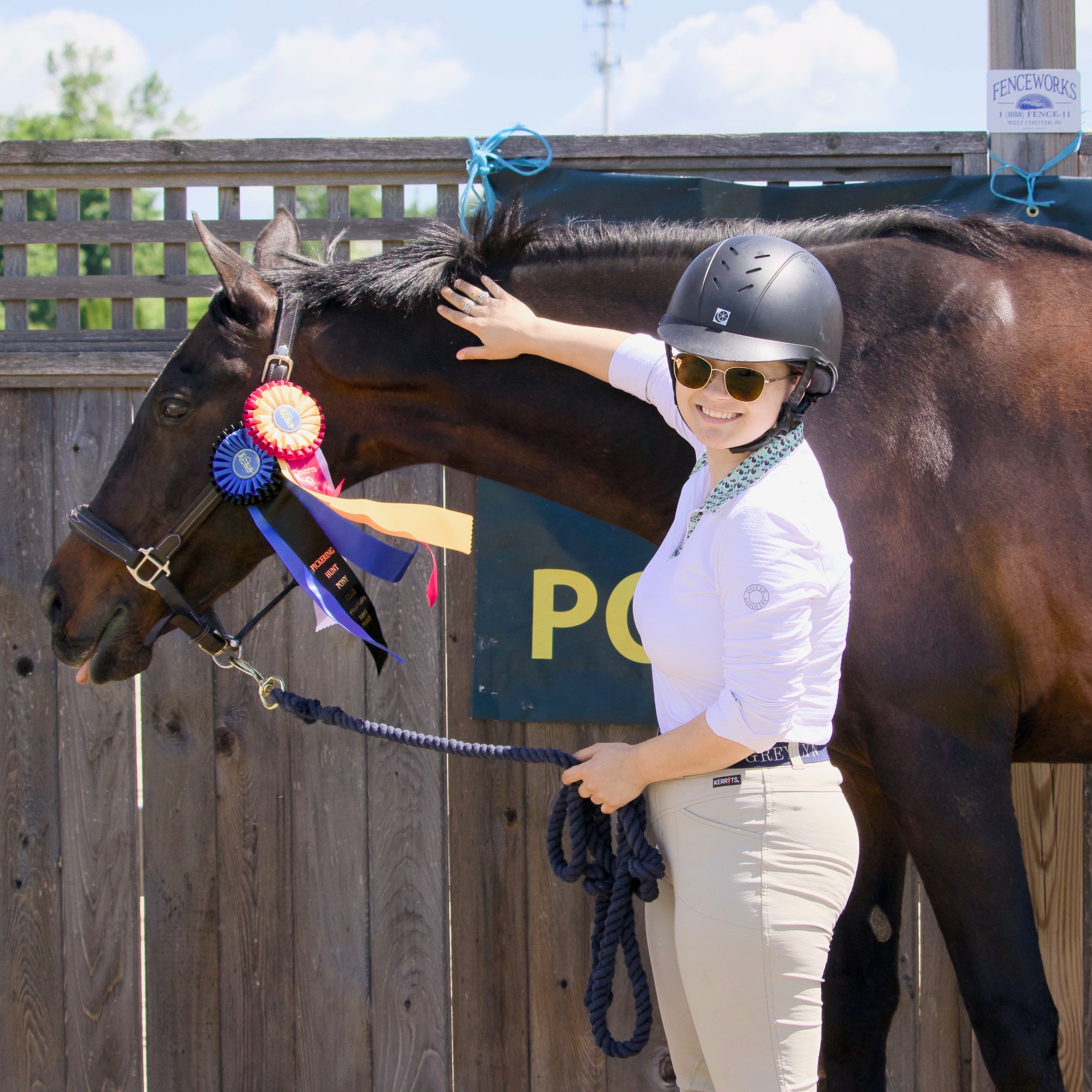 A person wearing a helmet and sunglasses petting a horse with a red and blue ribbon on its bridle, in an outdoor wooden stall.