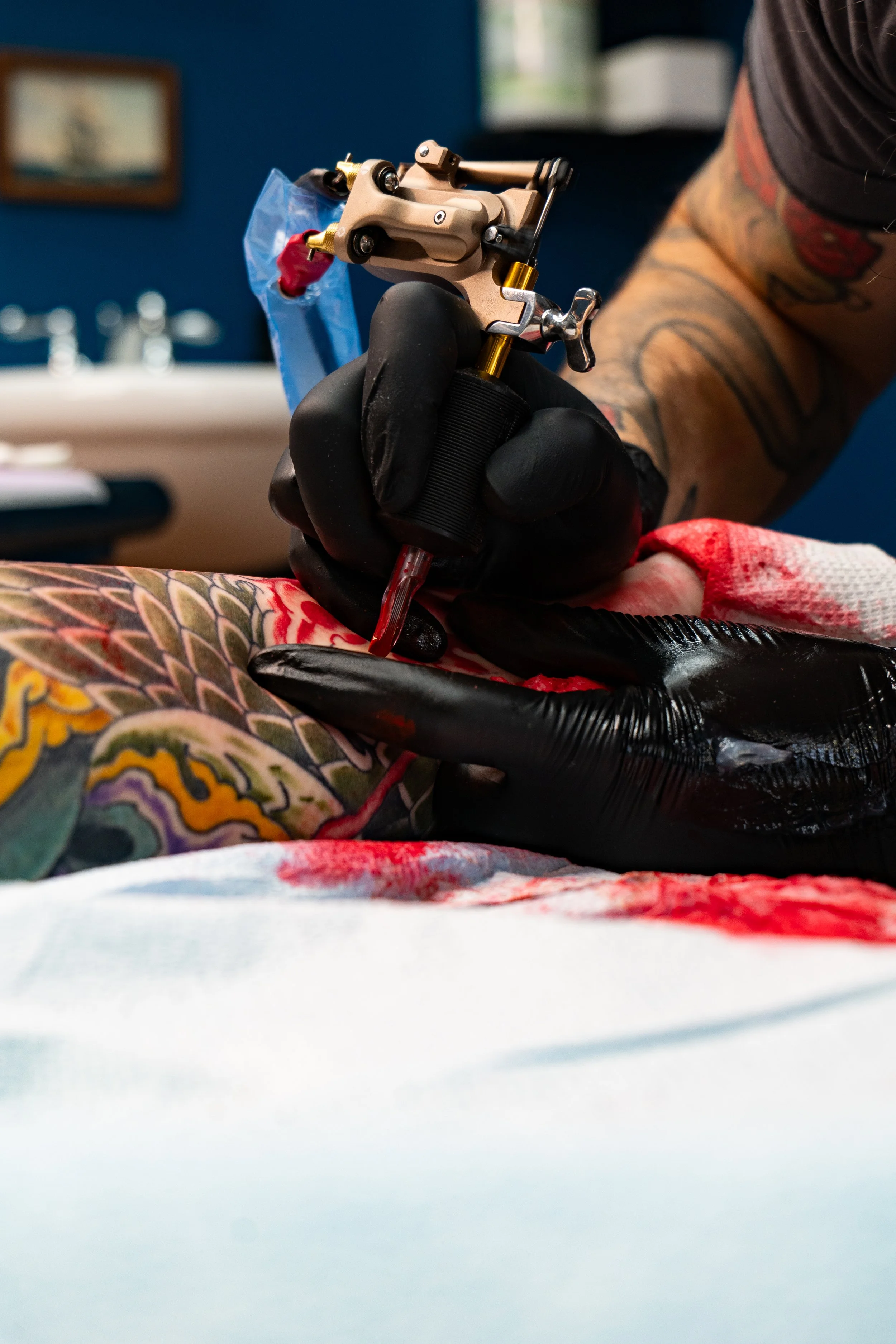 Tattoo artist wearing black gloves and holding a tattoo machine, working on a colorful tattoo on a person's arm.