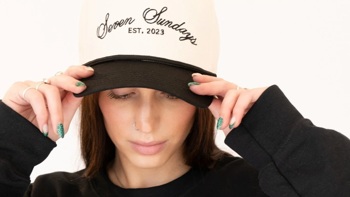 A woman with brown hair and a nose piercing adjusting a cap that says 'Born Sundays EST. 2023' against a plain white background.