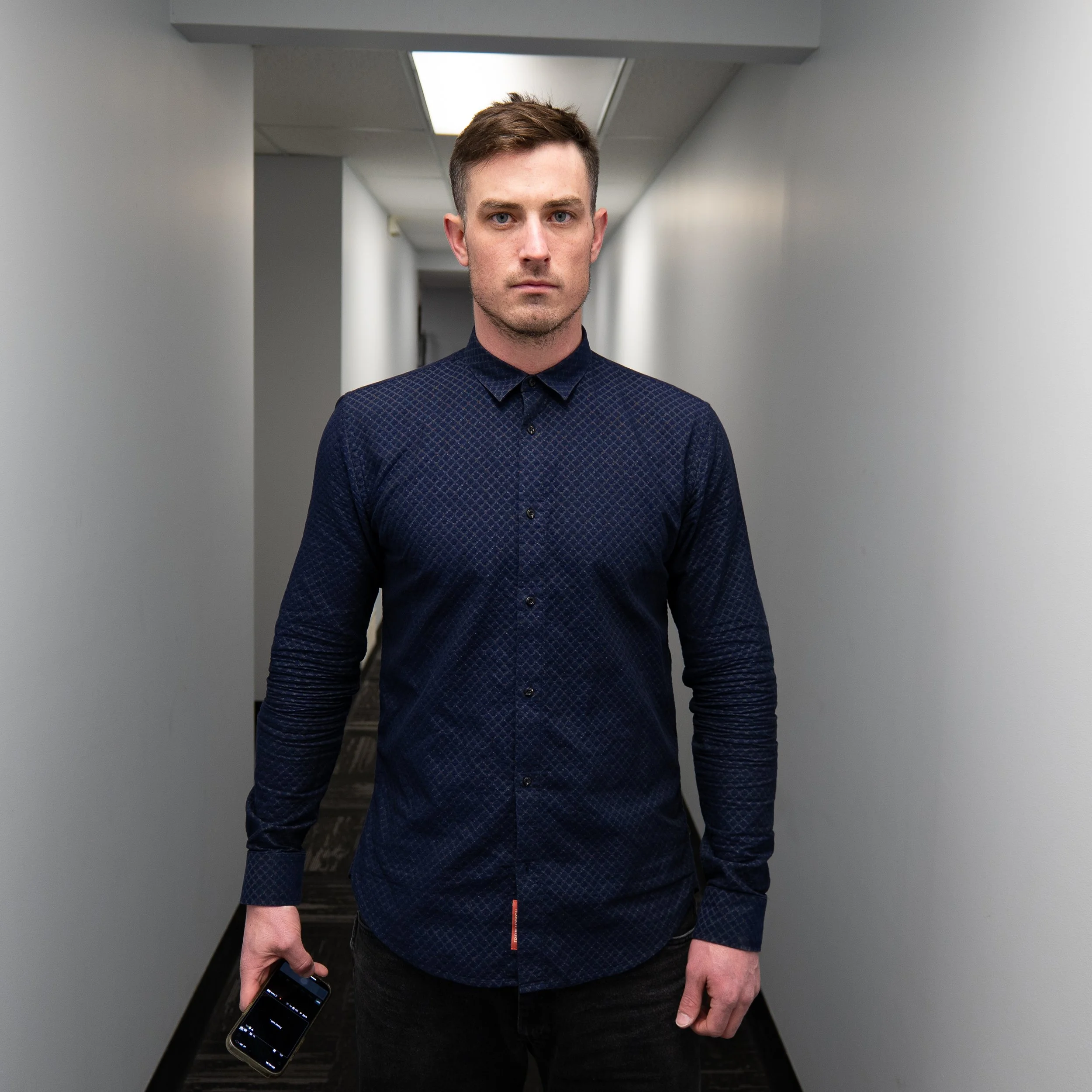 Cory Getz standing in a hallway, wearing a dark patterned button-up shirt and holding a smartphone in his right hand.