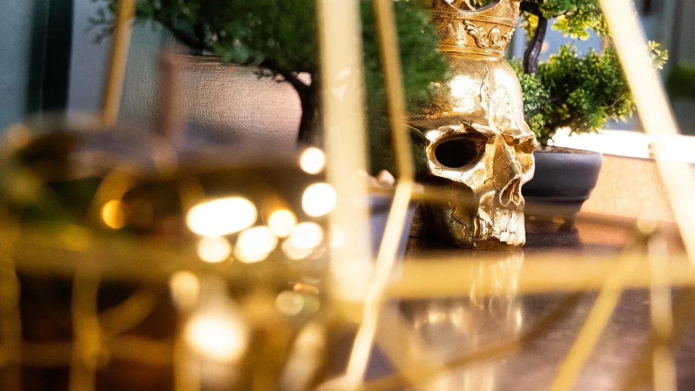 A gold-colored skull sculpture with plants in the background, positioned on a wooden surface, with soft lighting and blurred golden wire decorations in the foreground.