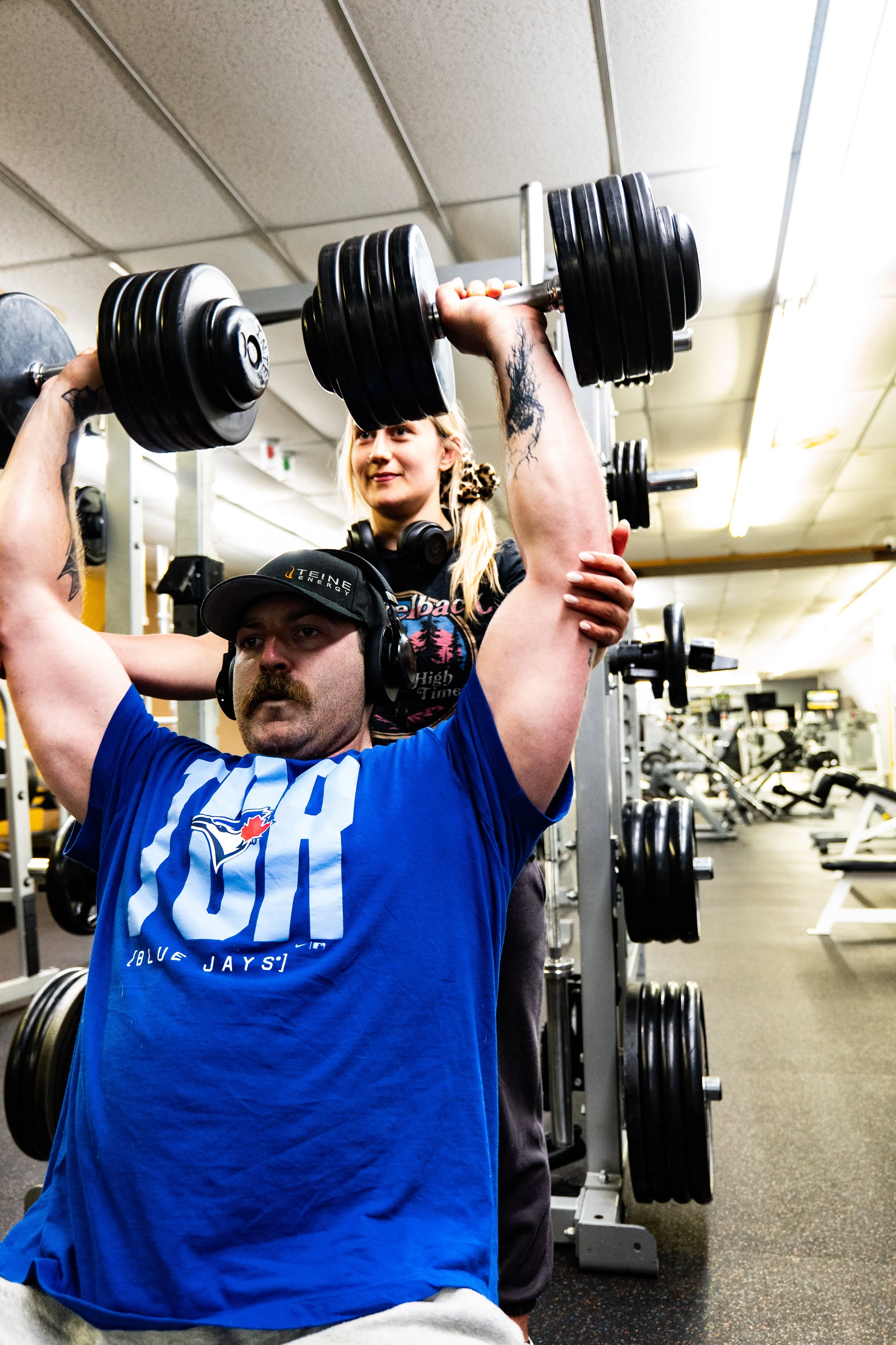A man in a blue sports shirt is lifting a dumbbell above his head in a gym. A woman in a black shirt and headphones is standing behind him, spotting him. The gym has various weightlifting equipment and a ceiling with fluorescent lights.