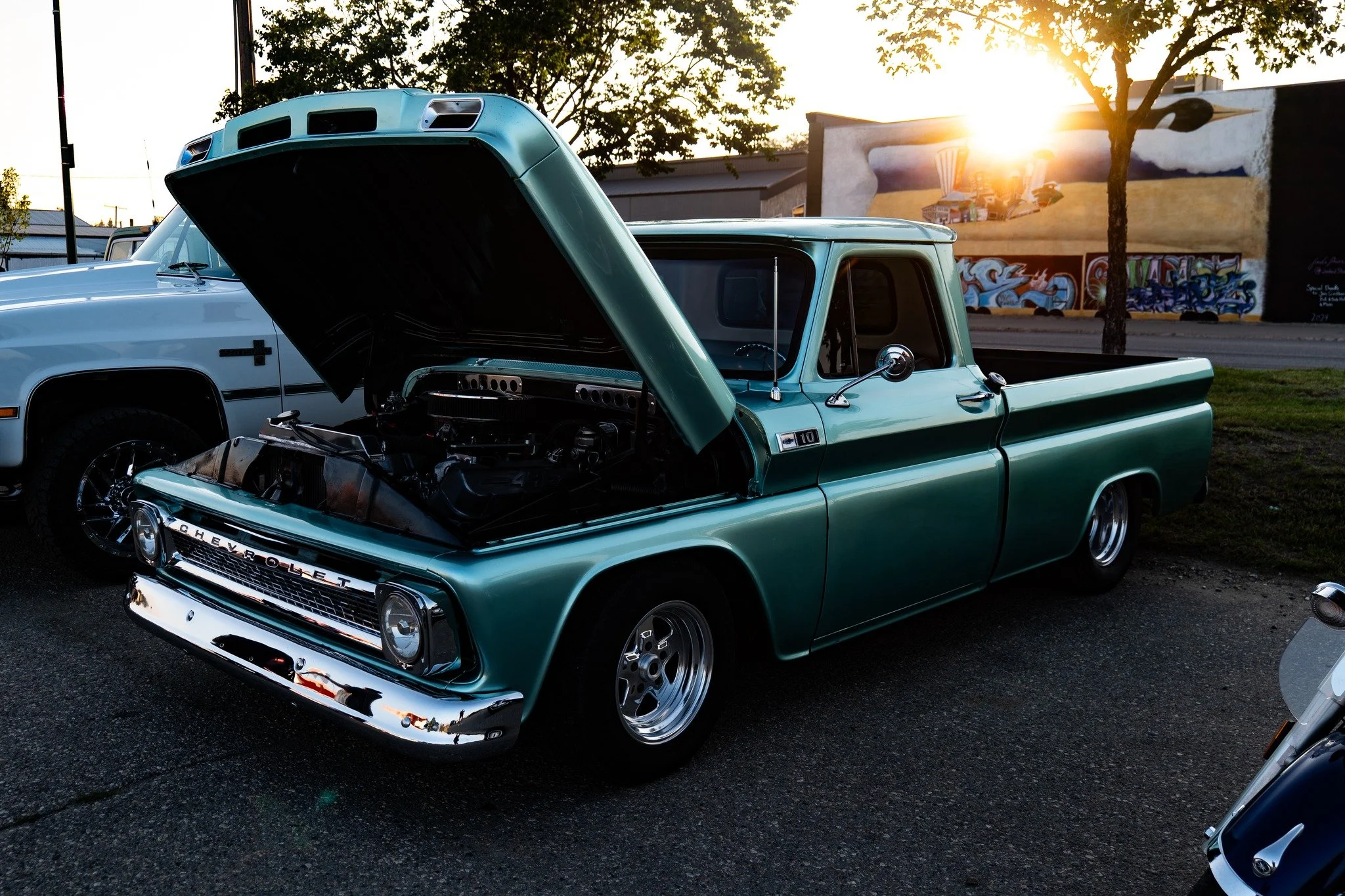 A vintage Chevrolet pickup truck with an open hood, parked outdoors during sunset. The truck is turquoise with chrome accents. In the background, there is a white vehicle, graffiti on a wall, and a tree, with the sun setting behind the scene.