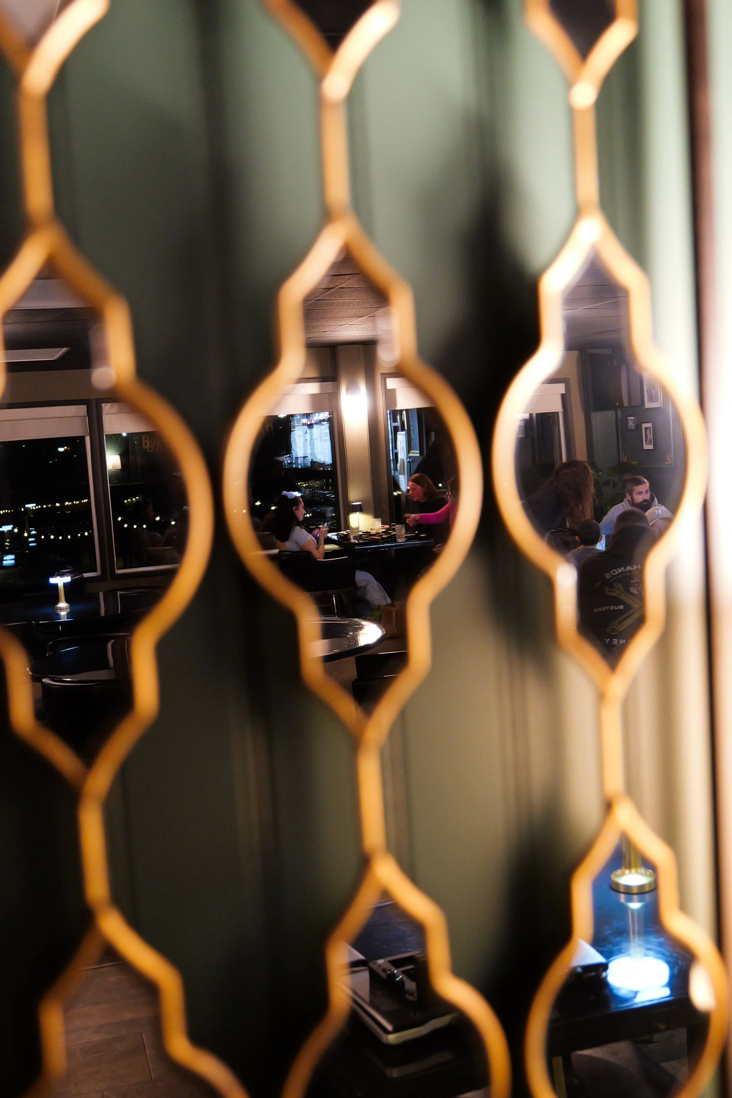 Interior of a restaurant or bar seen through a decorative metal screen with a honeycomb pattern. There are people sitting at tables, some drinking, with large windows showing city lights outside. The lighting inside is dim and cozy.