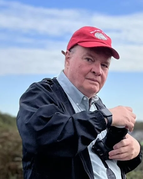 A man wearing a red baseball cap, a light blue collared shirt, and a dark jacket, holding a camera, outdoors under a partly cloudy sky.