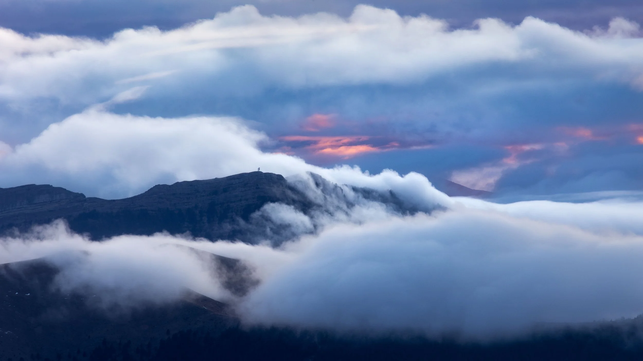 Mountain peaks surrounded by clouds and mist, with a hint of pink sky visible through the clouds.
