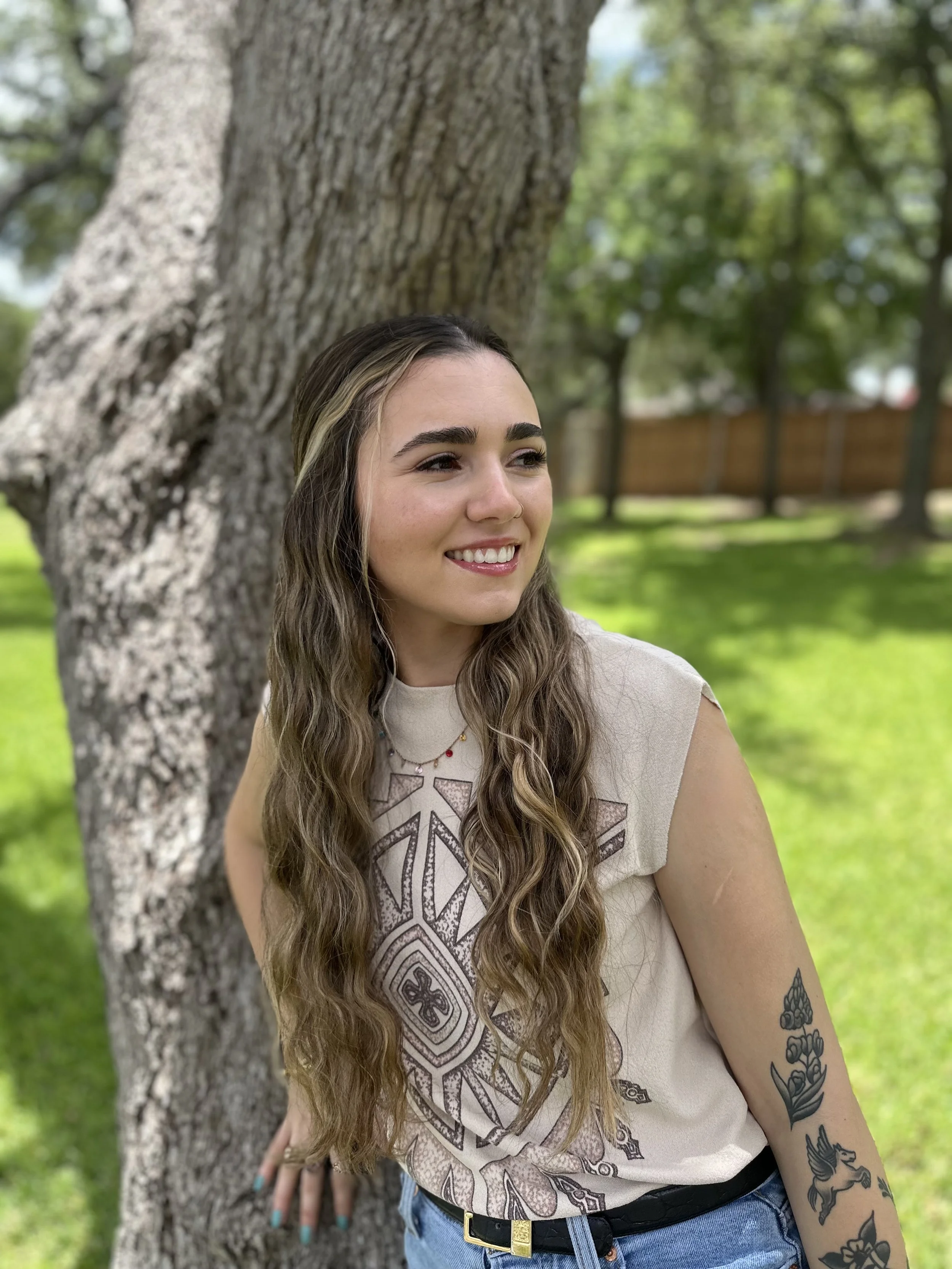 A young woman with long wavy hair, multiple tattoos on her arm, and a nose piercing, leaning against a tree outdoors in a park with a wooden fence and green trees in the background. She is smiling and wearing a beige T-shirt with a geometric design, blue jeans, and a black belt.