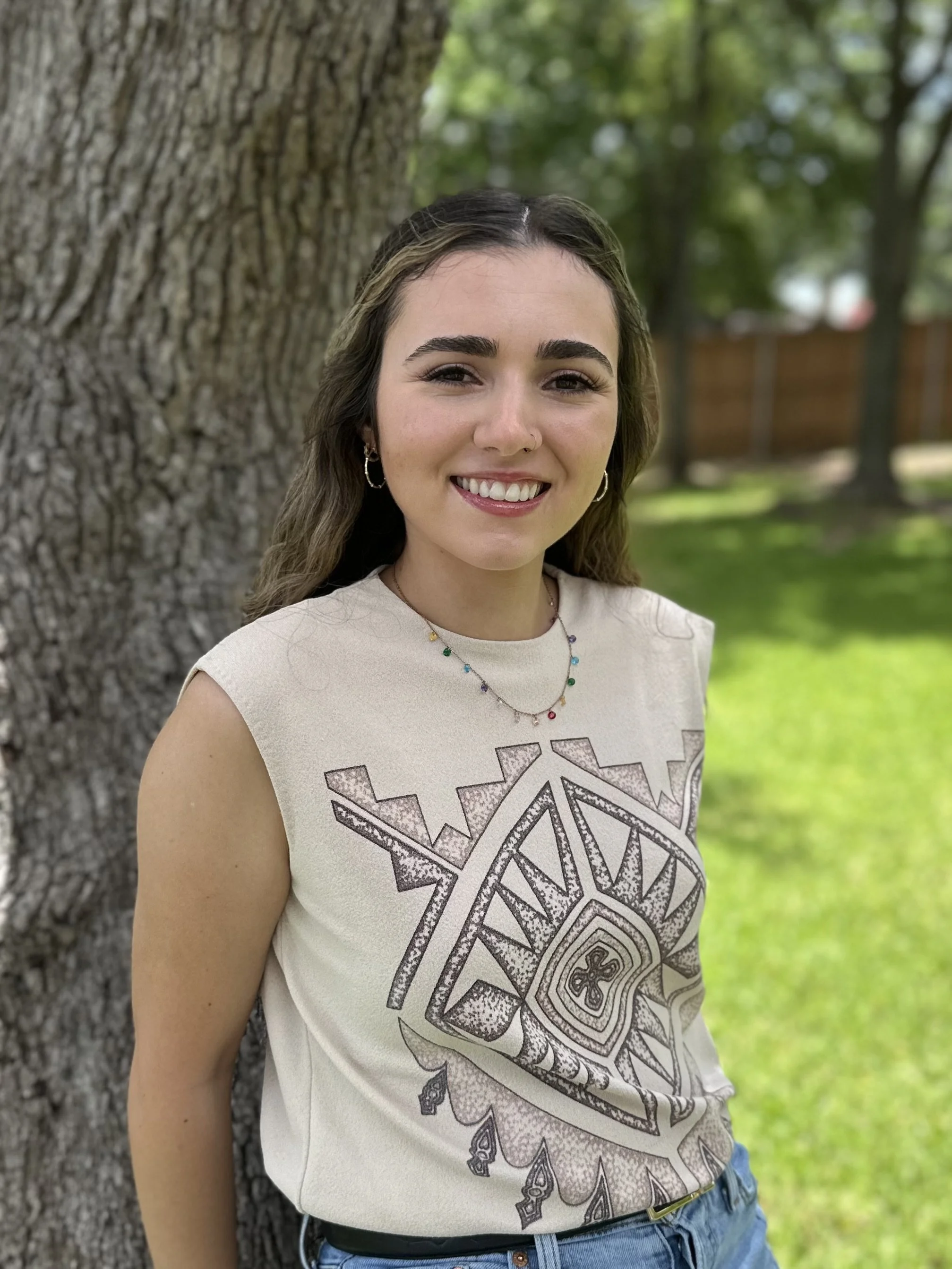 A young woman with brown hair and hoop earrings smiling outdoors near a large tree with a textured bark, wearing a beige graphic t-shirt and blue jeans, with a wooden fence and green trees in the background.
