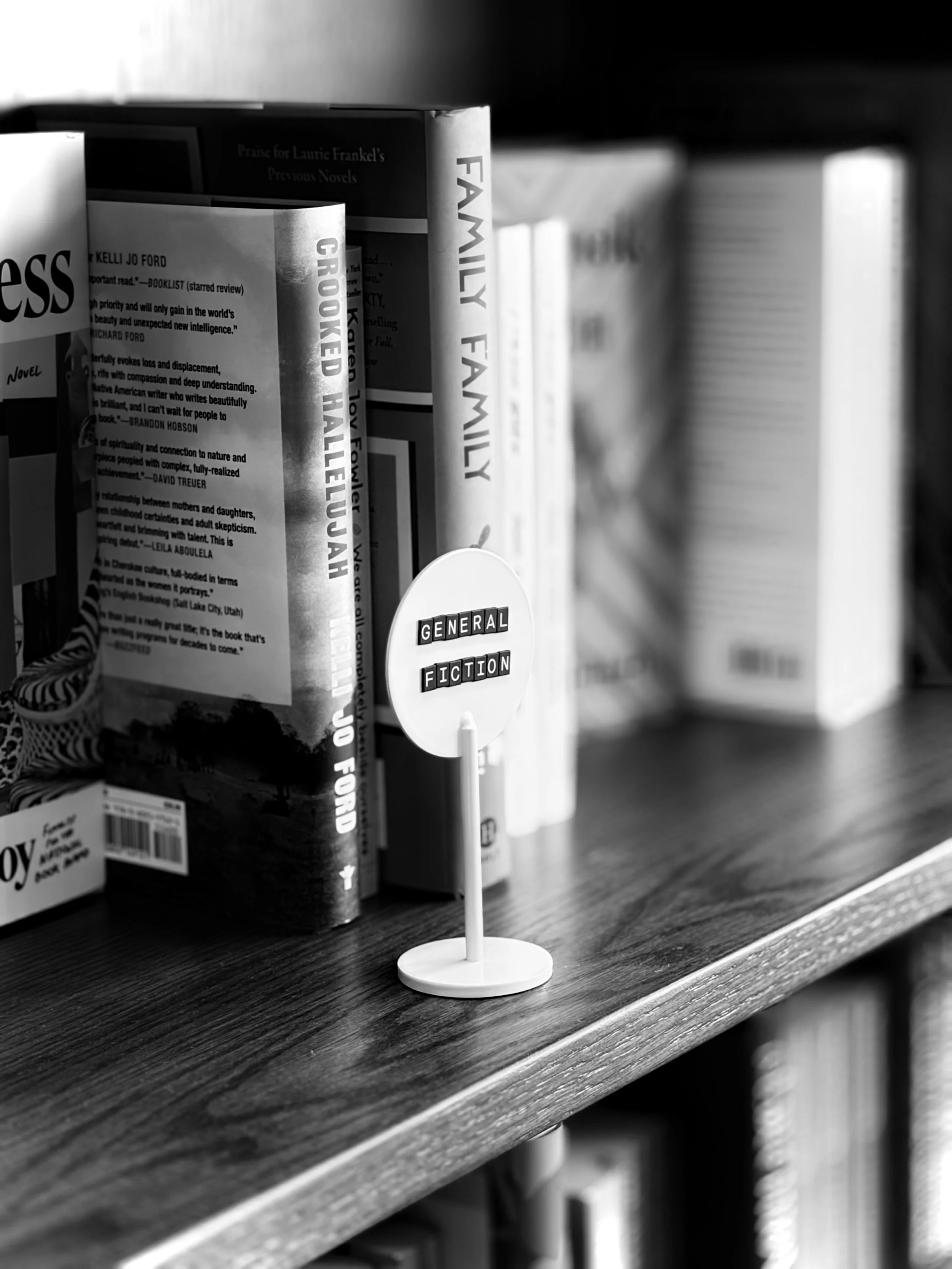 A black-and-white photo of a small white sign with the words 'GENERAL FICTION' on it, placed on a wooden shelf among books.