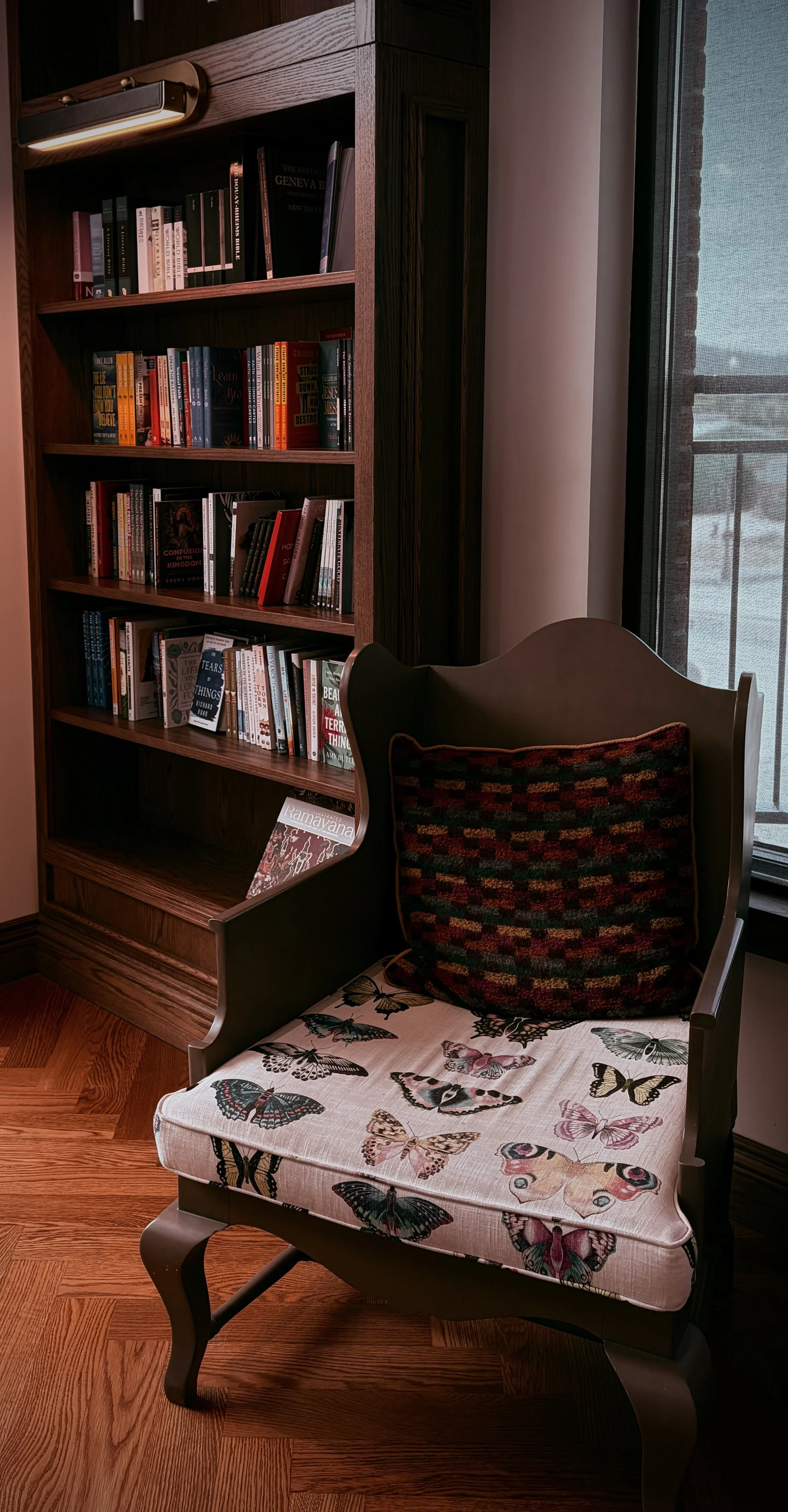 Wooden bookshelf filled with books next to a window with a screen, partially visible, and a colorful armchair with a butterfly-patterned cushion.