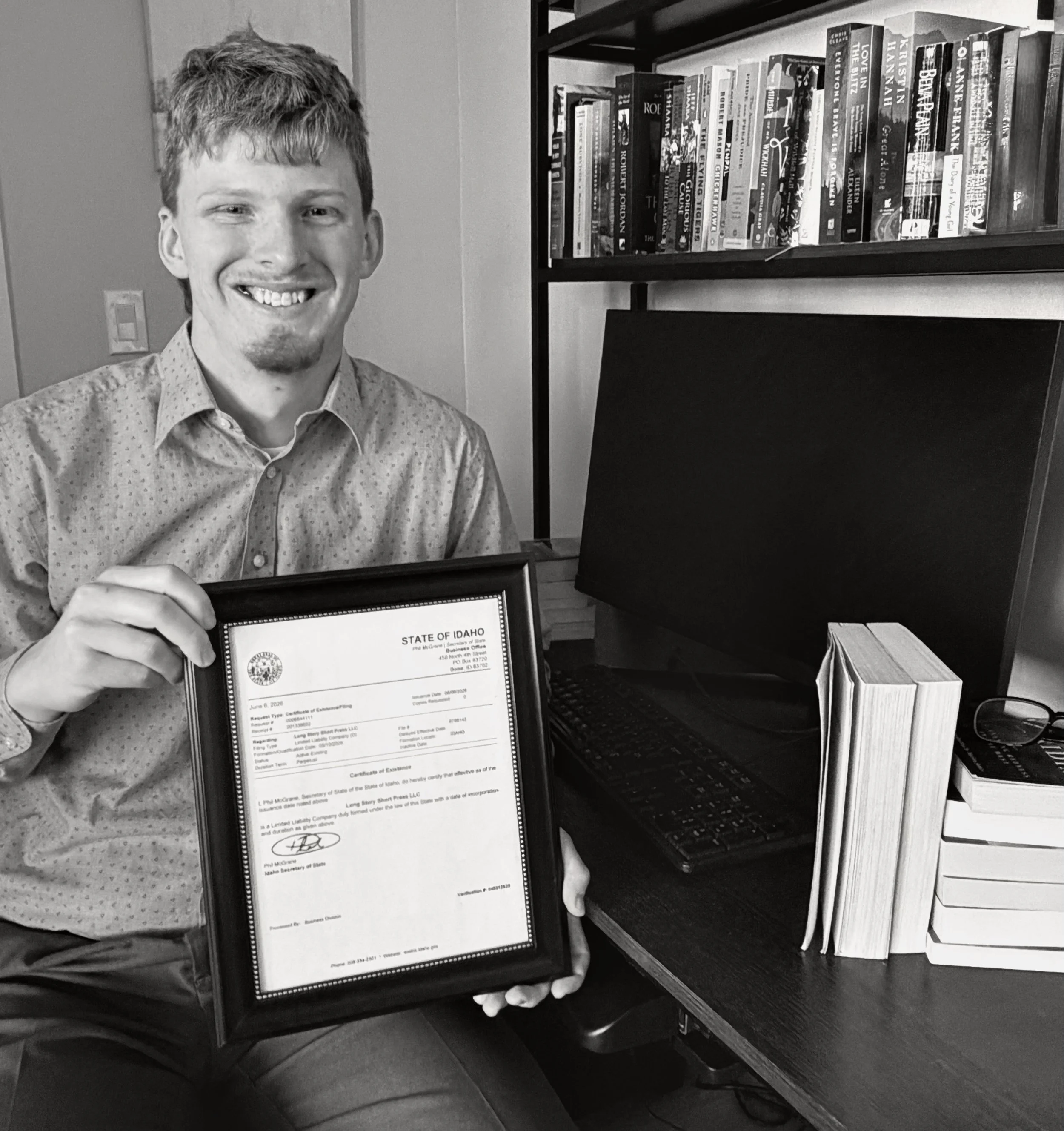A smiling man with short, curly hair wearing a button-up shirt holding a framed certificate. He is standing next to a bookshelf filled with various books and a computer monitor. The setting appears to be an indoor office or study.