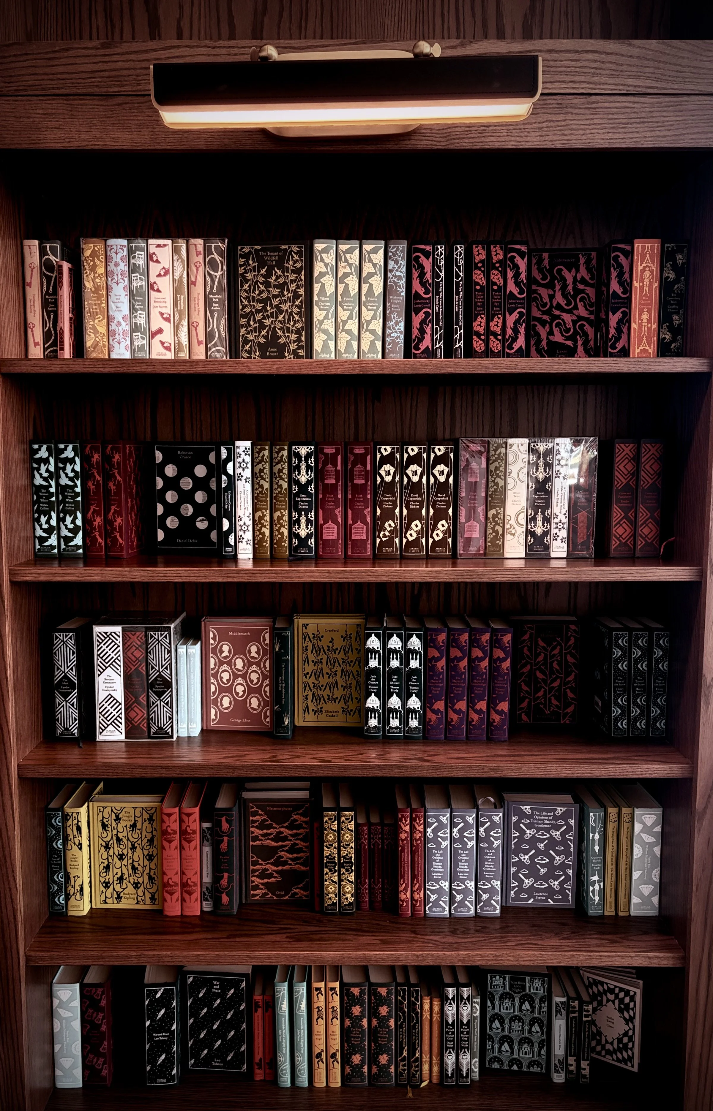 A wooden bookshelf filled with various books organized by color, including black, white, red, yellow, and gray, with a light above illuminating the top shelf.