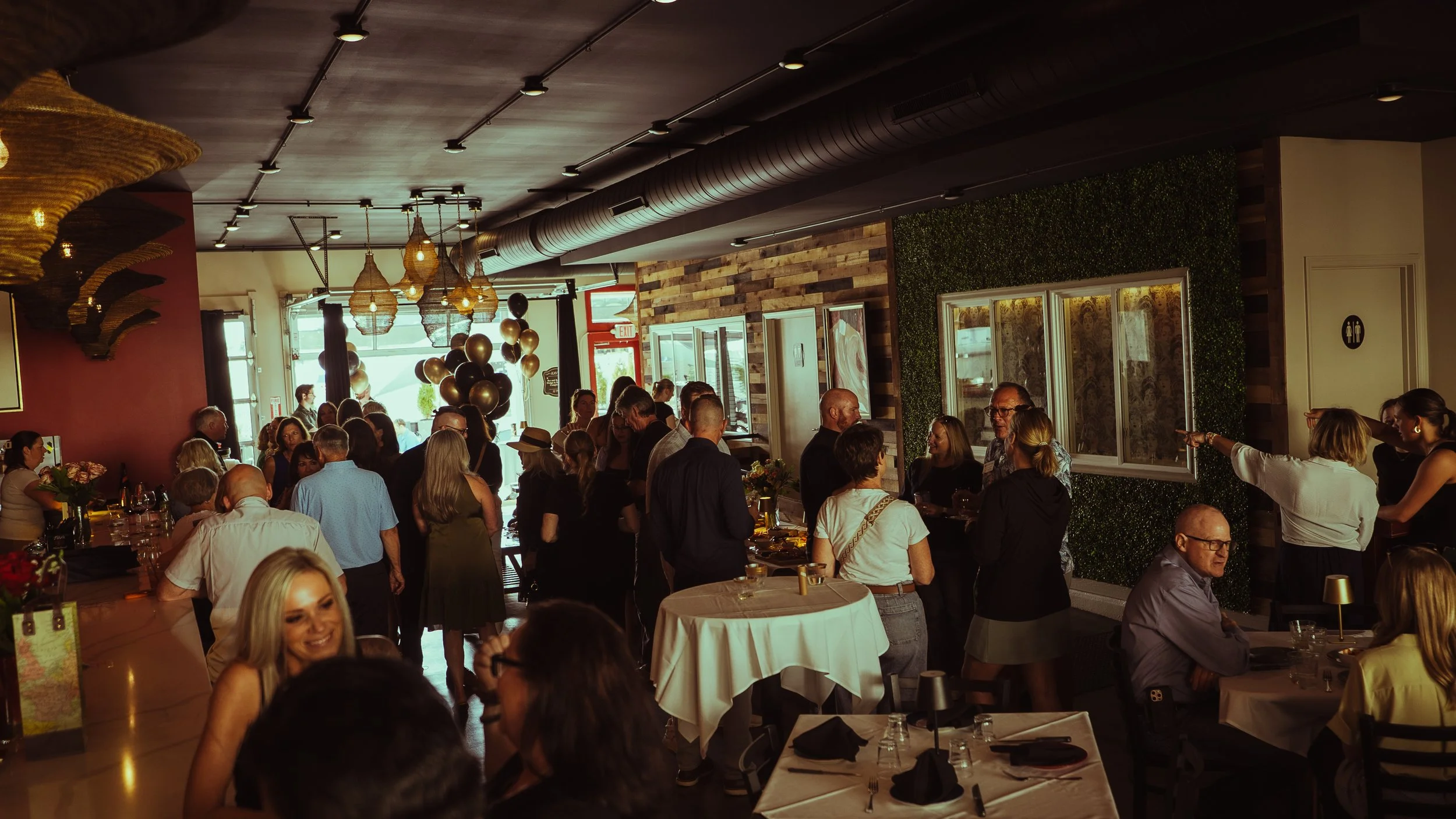 Crowd of people gathered inside a restaurant with modern decor, hanging lights, and a green wall, during a social event or celebration.