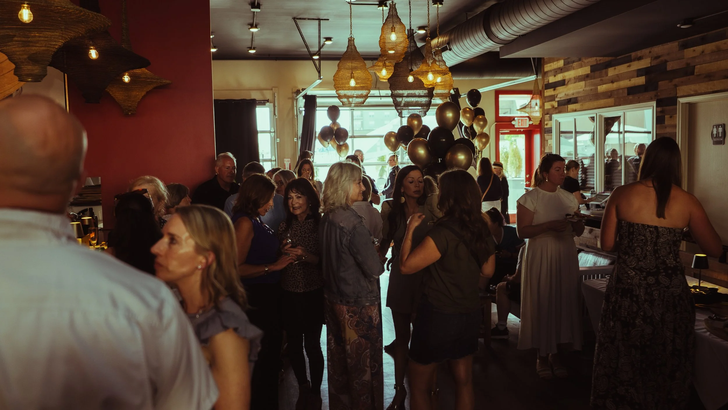 People gathered inside a restaurant or event space decorated with black and gold balloons and warm hanging lights.