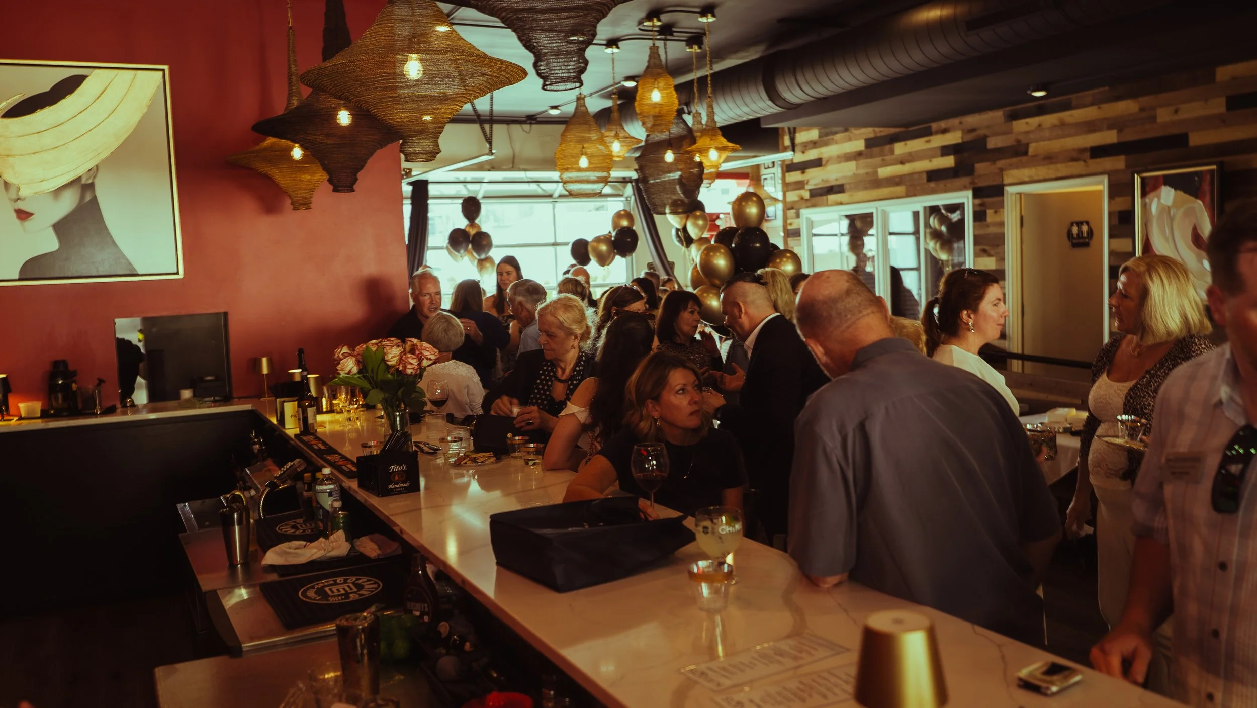 A crowded bar or restaurant with people socializing, some holding drinks, in a warmly lit interior with balloons and artistic wall decor.