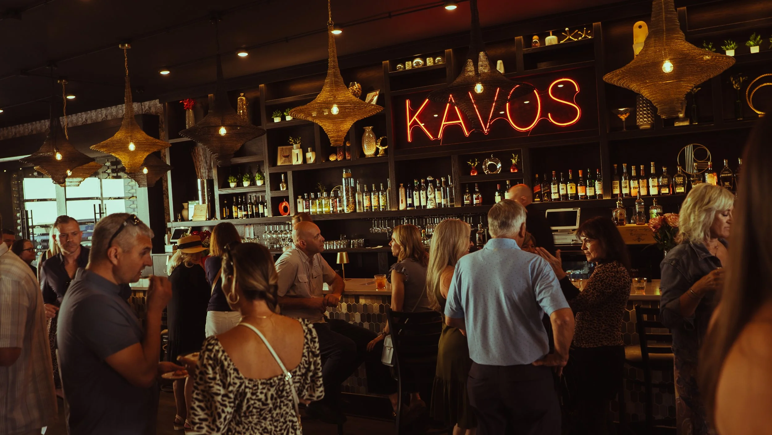 A lively bar scene with people socializing, some holding drinks, inside a dimly lit bar named Kavos with a neon sign, stylish lighting fixtures, and shelves filled with bottles and decor.