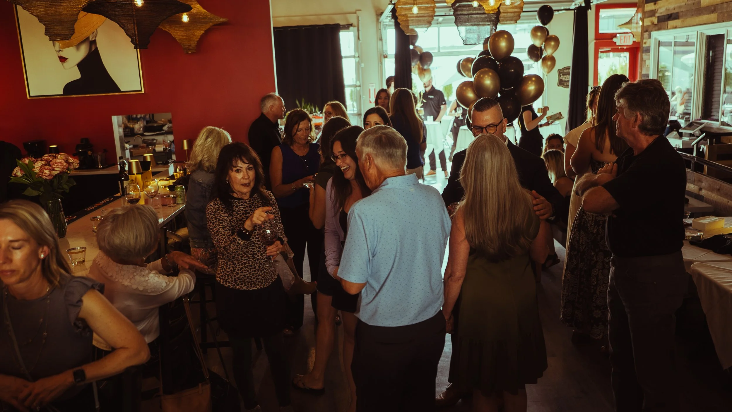 A crowded indoor party with people socializing, balloons, and decorations.