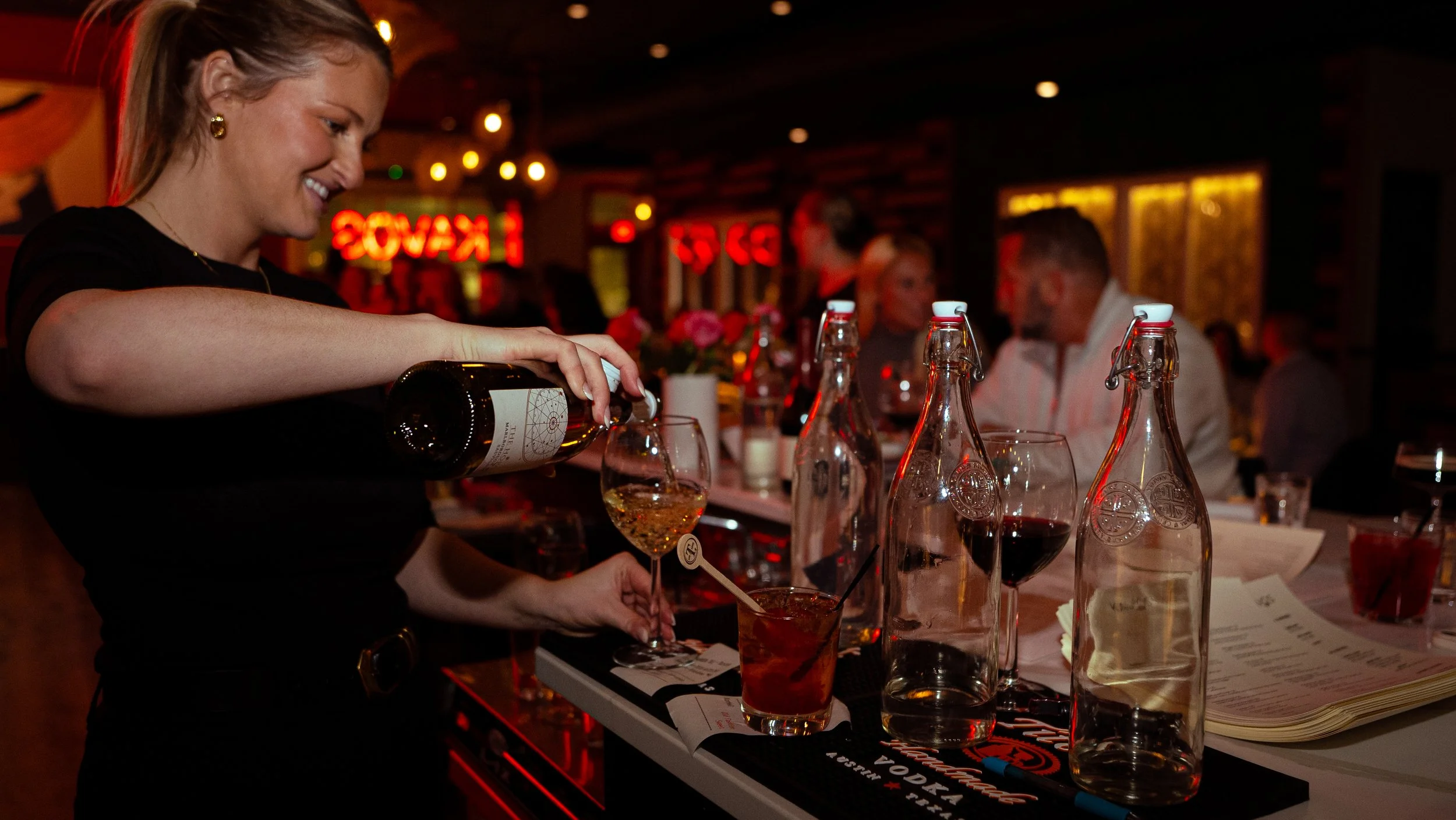 A smiling woman is pouring a drink into a glass at a bar. There are multiple glass bottles and a menu on the bar counter. In the background, other people are sitting and chatting in a warmly lit restaurant or bar with neon signs.