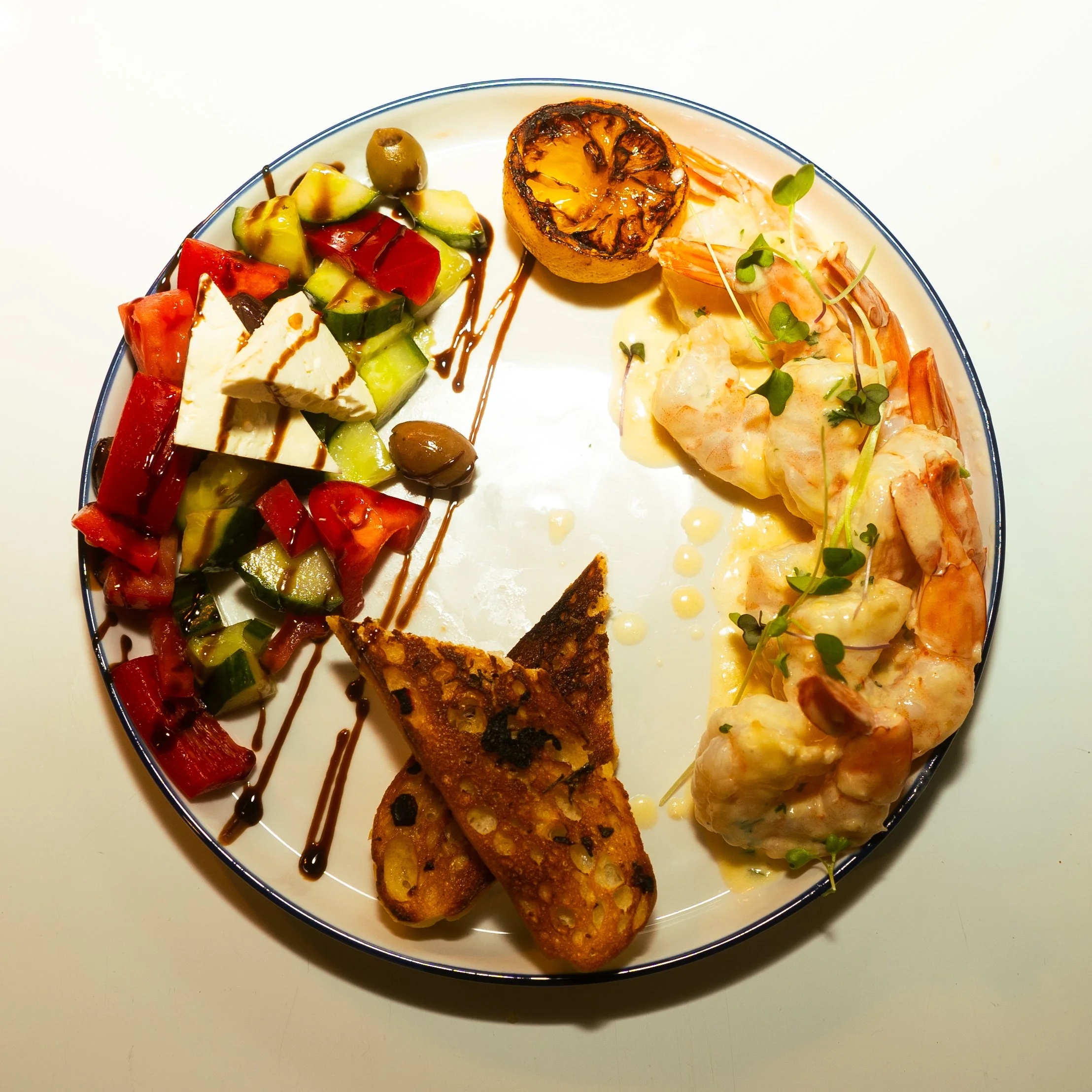 A white plate with a rim contains a variety of foods, including grilled lemon half, seafood, chopped vegetables, and two slices of toasted bread, all garnished with microgreens and drizzled with sauce.