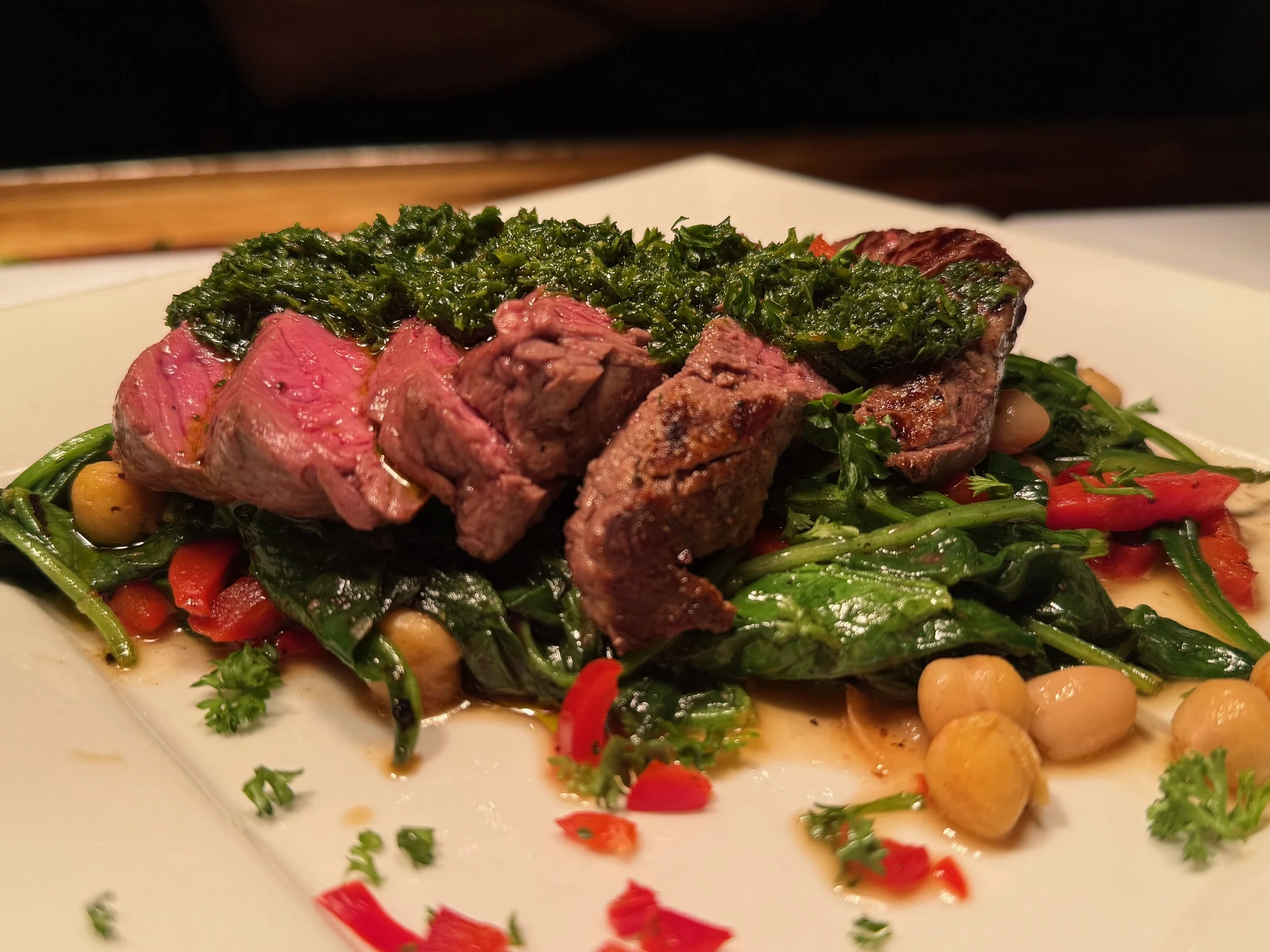 Sliced medium-rare steak topped with sautéed greens garnished with red pepper flakes, served on a white plate with chickpeas and vegetables.