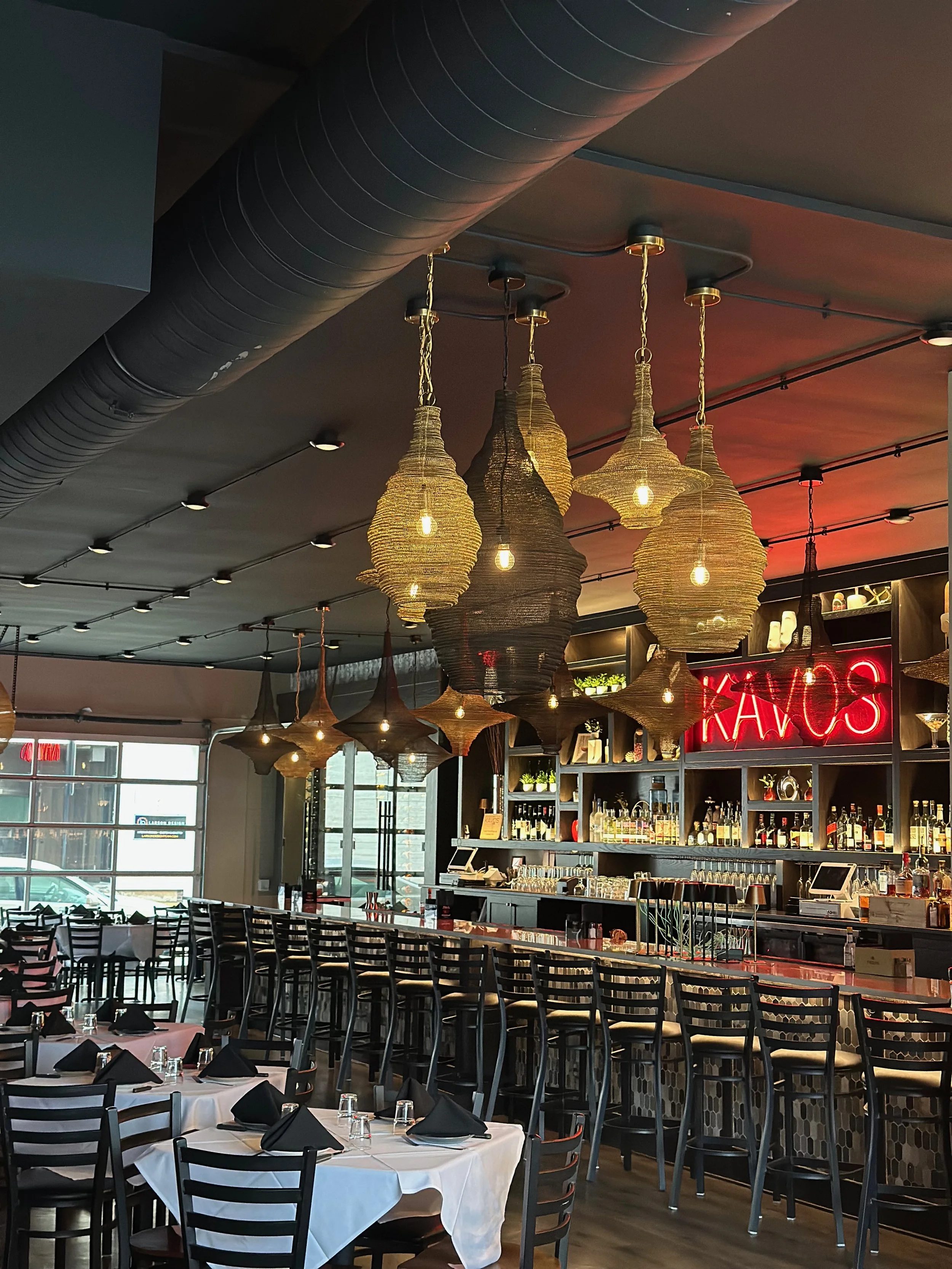 Interior of a restaurant with tables set for dining, featuring black chairs, white tablecloths, and black napkins. Decorative woven pendant lights hang from the ceiling, and a bar area with shelves of bottles and glasses is visible in the background. Neon red sign reading 'Kavos' is also present.