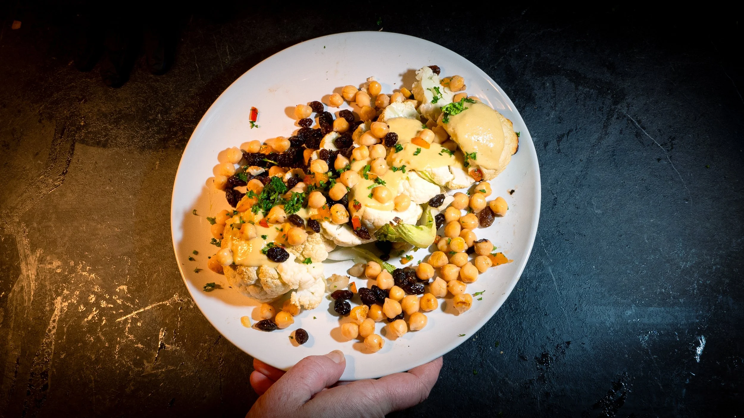 A white plate of roasted cauliflower topped with a creamy yellow sauce, garnished with chopped parsley, surrounded by roasted chickpeas and black beans on a dark surface.