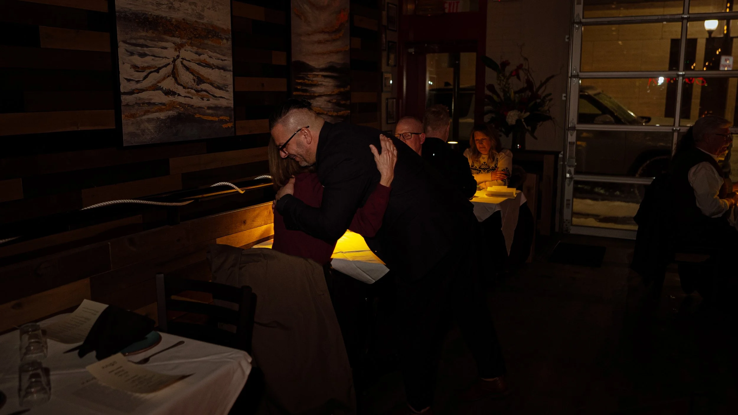 Two people embracing in a dimly lit restaurant. Other diners are visible in the background, and large windows reveal the outside at night.