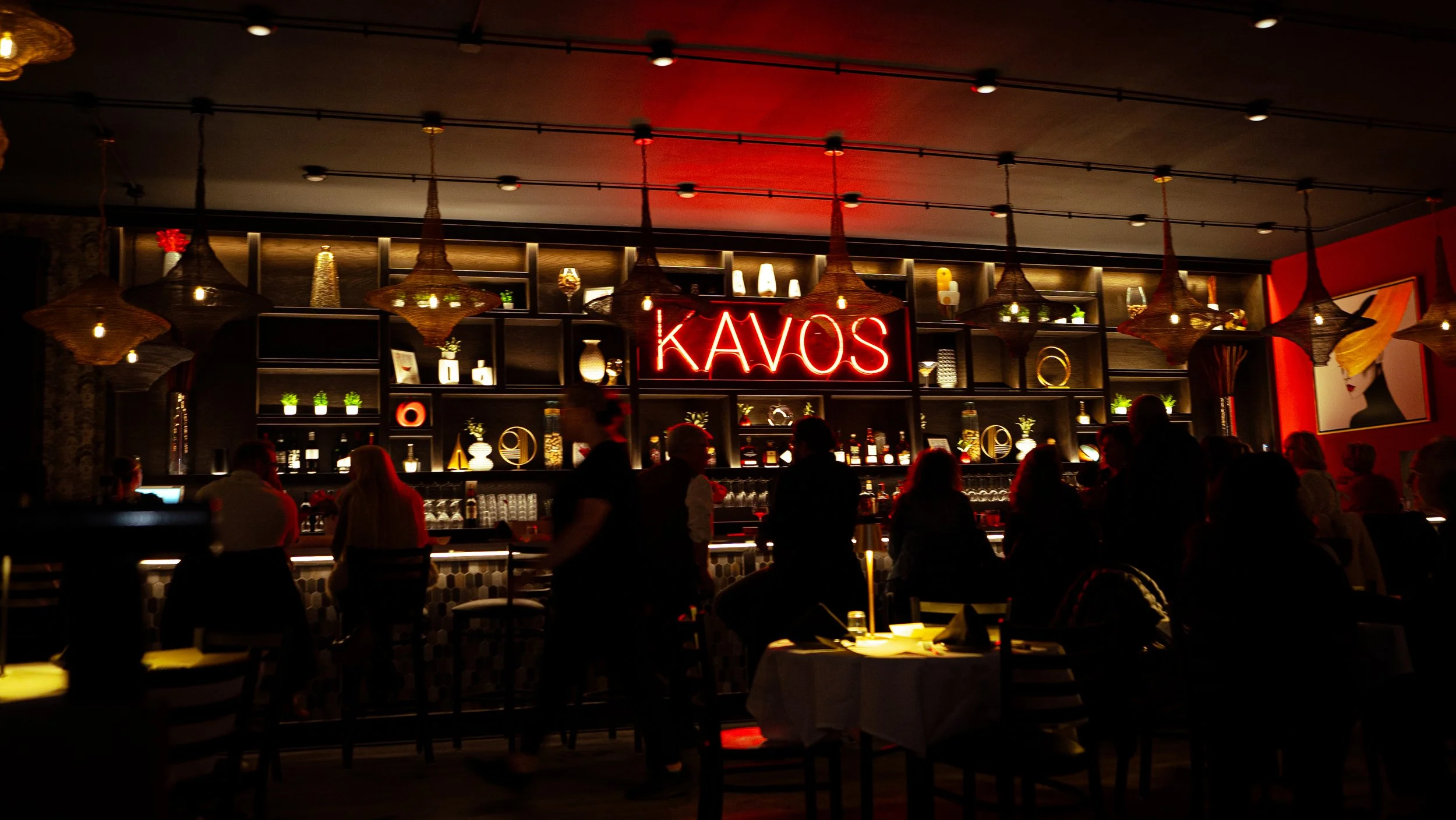 A cozy, dimly lit bar with a shelf filled with decor, glassware, and bottles, and a neon sign that says 'KAVOS'. There are people sitting and standing around, enjoying drinks.