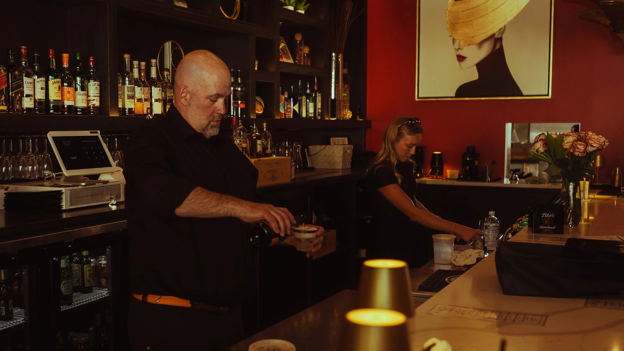Barman pouring a drink at a bar with alcohol bottles, a woman preparing a cocktail, and decorative flowers in a dimly lit, stylish bar.