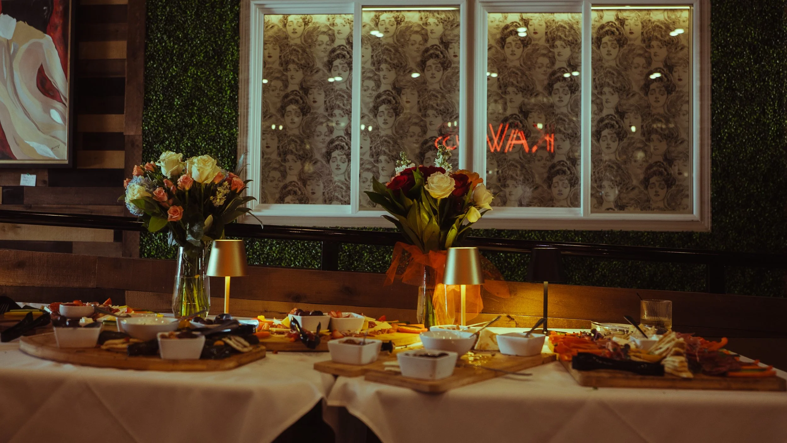 A restaurant or cafe interior with a buffet table filled with various dishes and bowls. Two vases with fresh flowers sit on the table. Behind the table, there is a large window with a retro wallpaper pattern of woman's faces, and a neon sign that reads 'OPEN'. The wall beside the window features a large portrait painting.