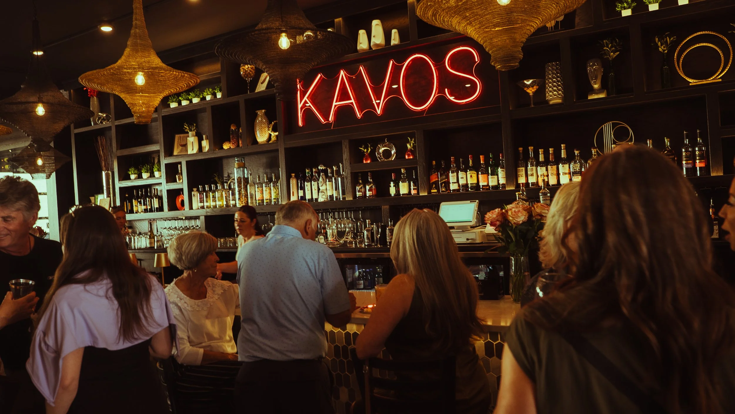 A busy bar with people socializing and a neon sign that reads 'KAOS' above the shelves of liquor bottles.