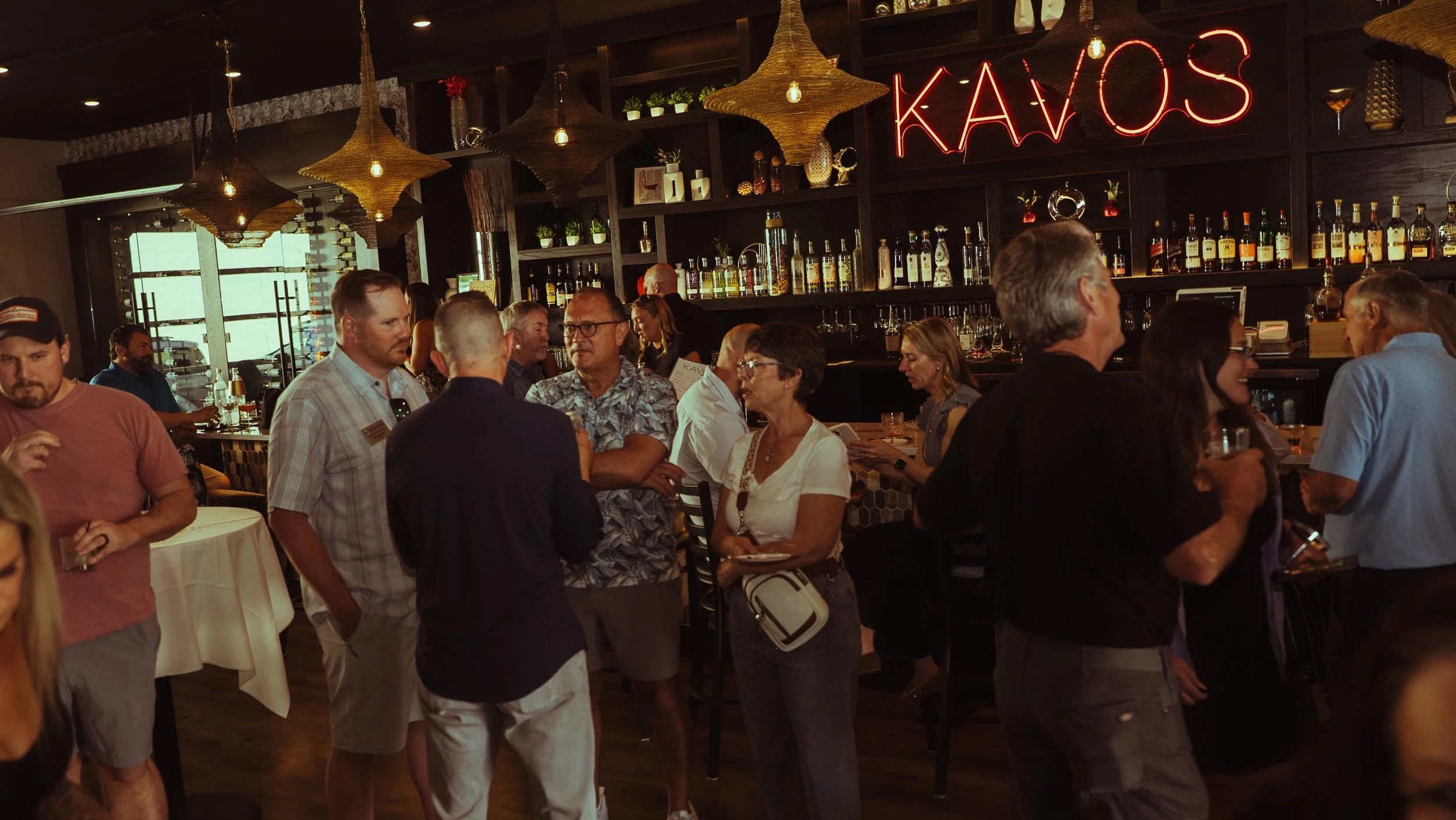 People socializing and mingling inside a bar or restaurant with a bar counter, bottles, and glasses in the background, and a neon sign that reads 'KAVOS'.