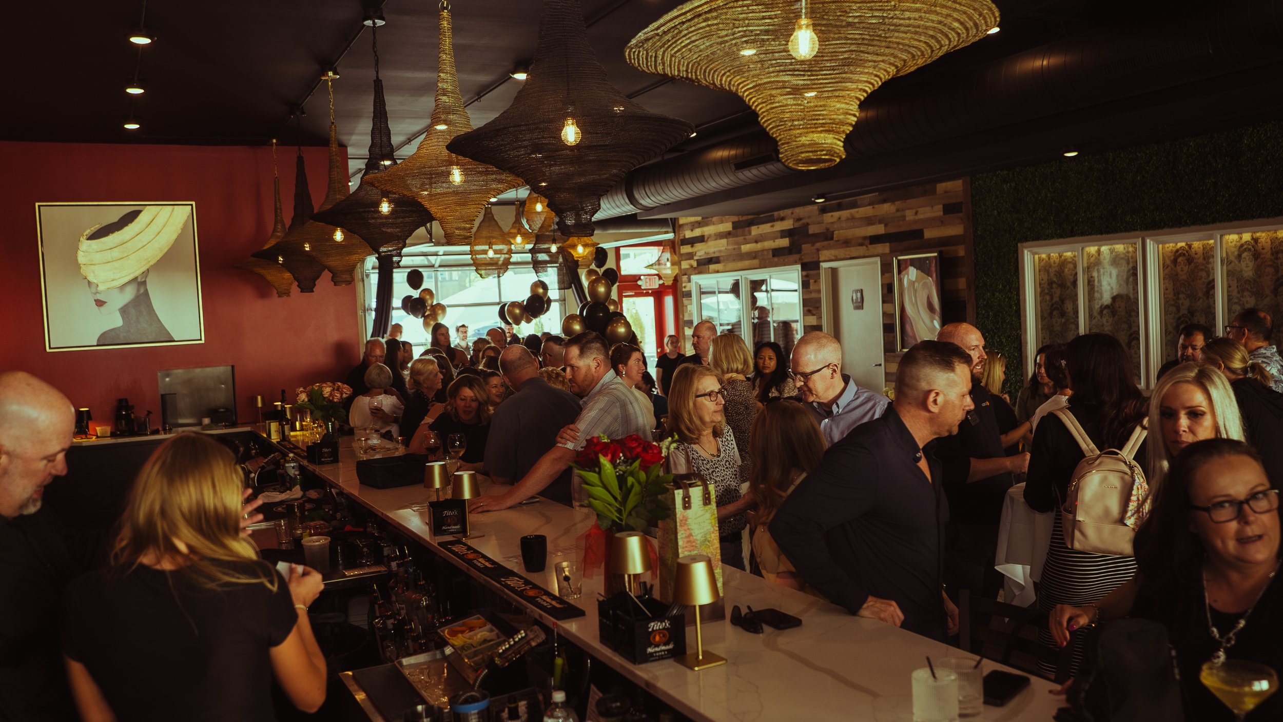 Crowded bar with people socializing inside a restaurant or bar, decorated with modern art paintings and unique hanging light fixtures.