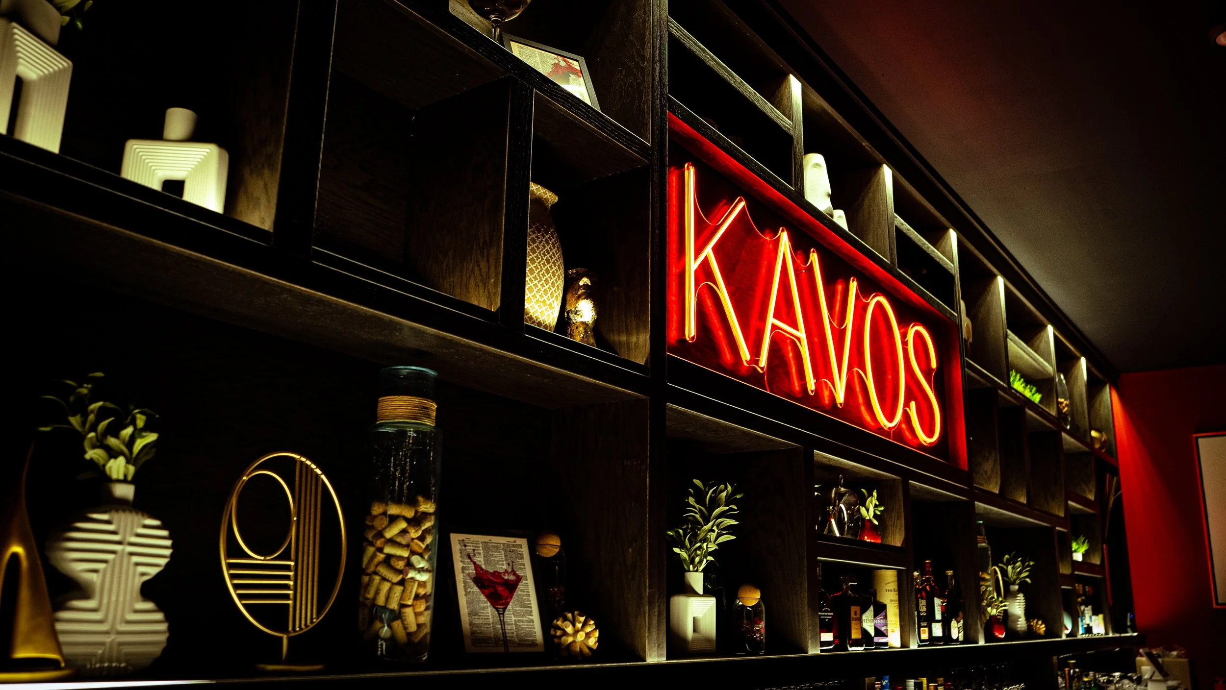 A dark wooden shelf with decorative items, including a white plant pot, a tall glass jar filled with corks, framed art, and a neon red sign reading 'NAYO'S' in a bar or restaurant setting.
