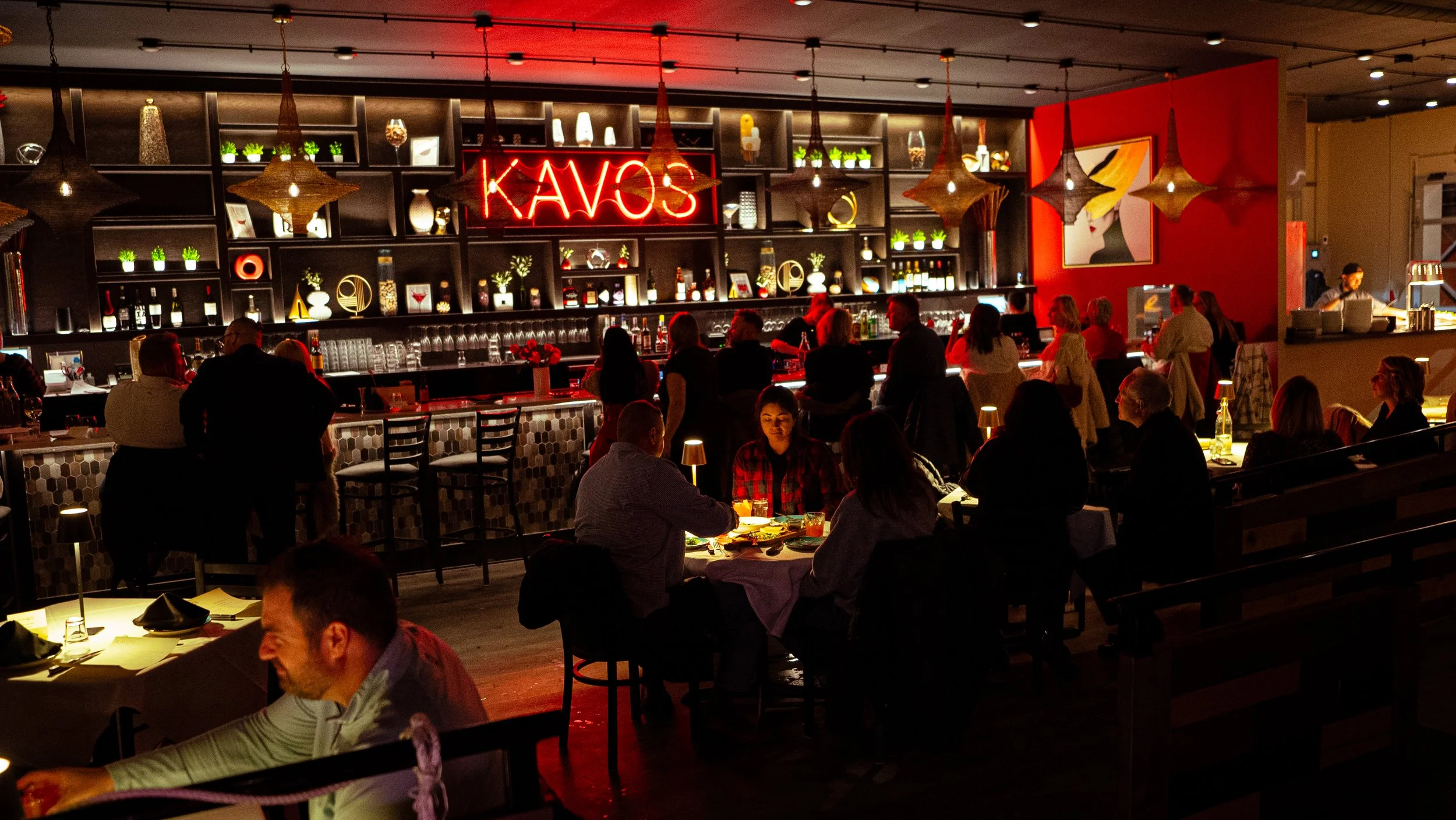 A busy restaurant with customers seated at tables and a bar area with a neon sign that reads 'KAVOS'. The decor includes hanging light fixtures, shelves with decorative items, and artwork on the walls.