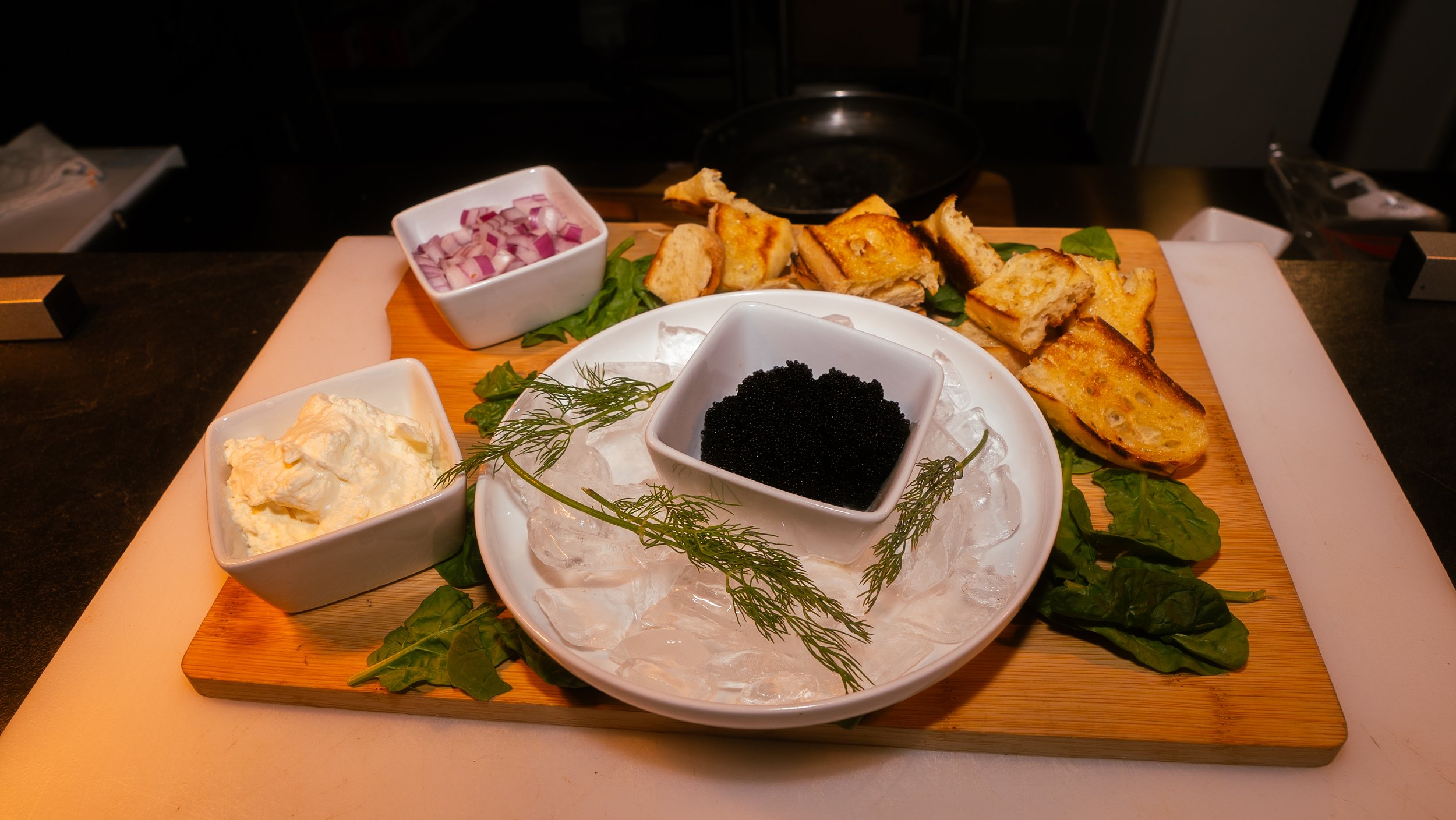 A wooden platter with various appetizers including scallion, potato pancake slices, caviar, egg salad, and chopped red onions.