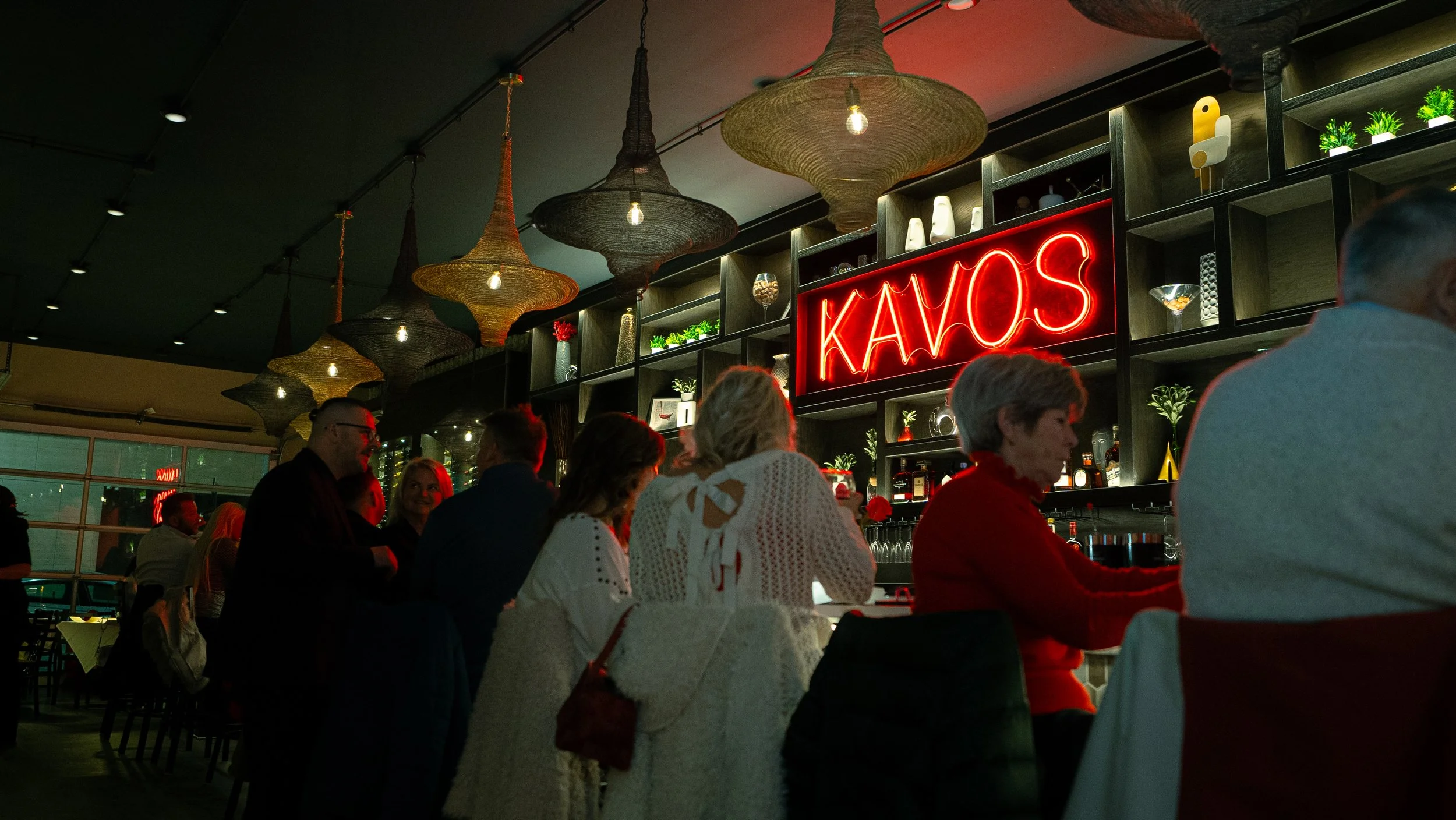 People dining inside a restaurant with a neon sign reading 'KAOS' behind the bar, hanging woven pendant lights, and various decorative items on shelves.