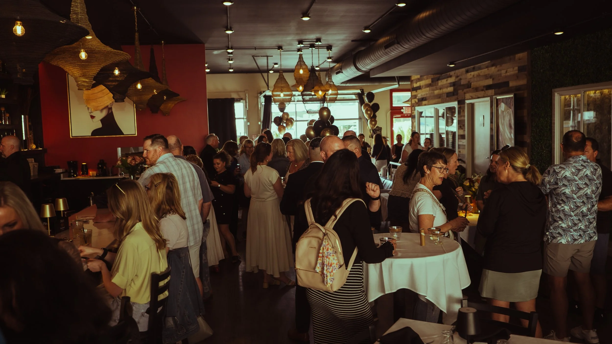 A busy indoor event with many people socializing. The decor includes hanging woven lampshades, balloon decorations, contemporary artwork on the walls, and warm lighting, creating a lively atmosphere.