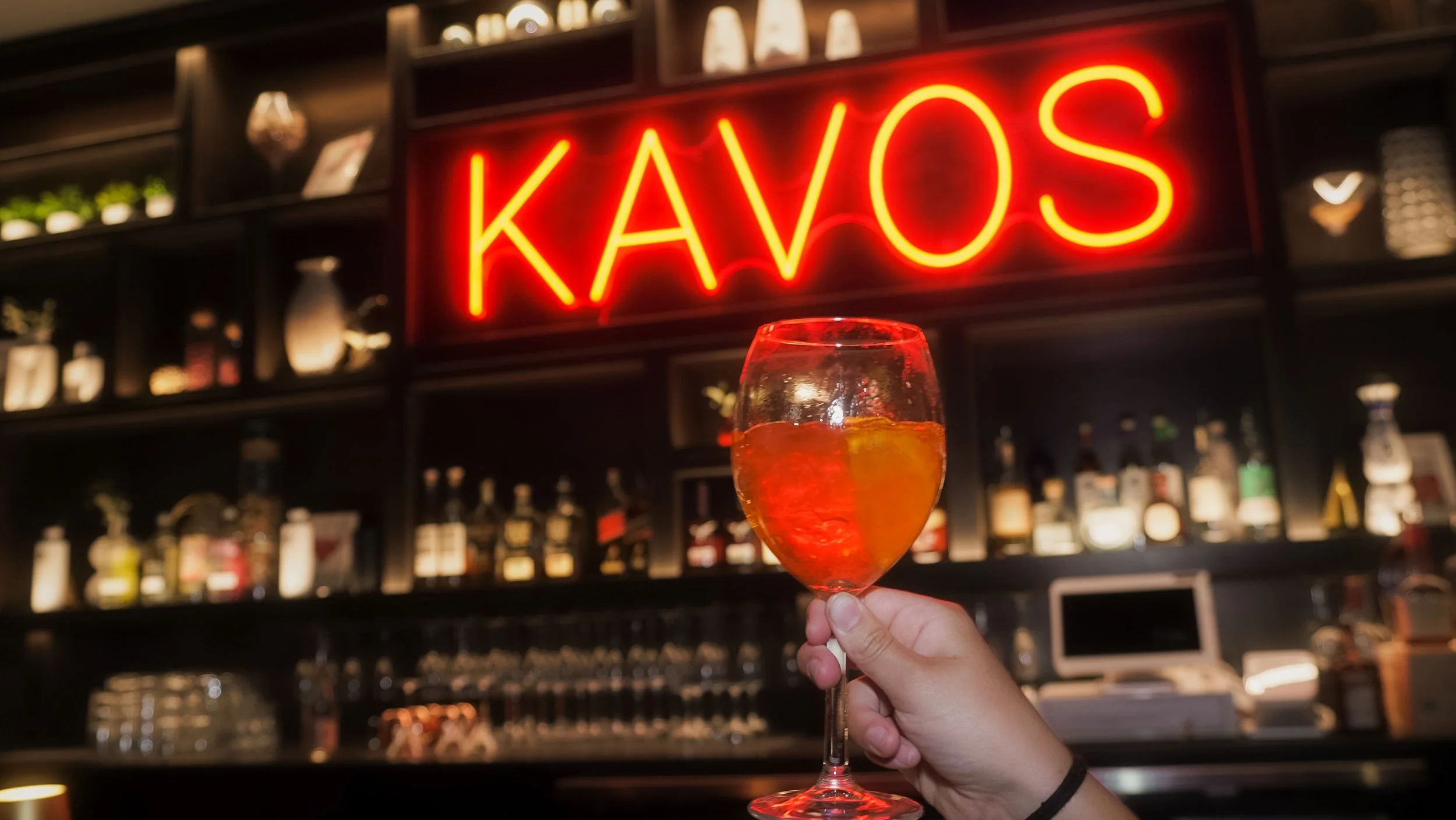 A hand holding a wine glass with a pink drink and an orange slice in front of a bar with a neon sign that says "KAVOS".