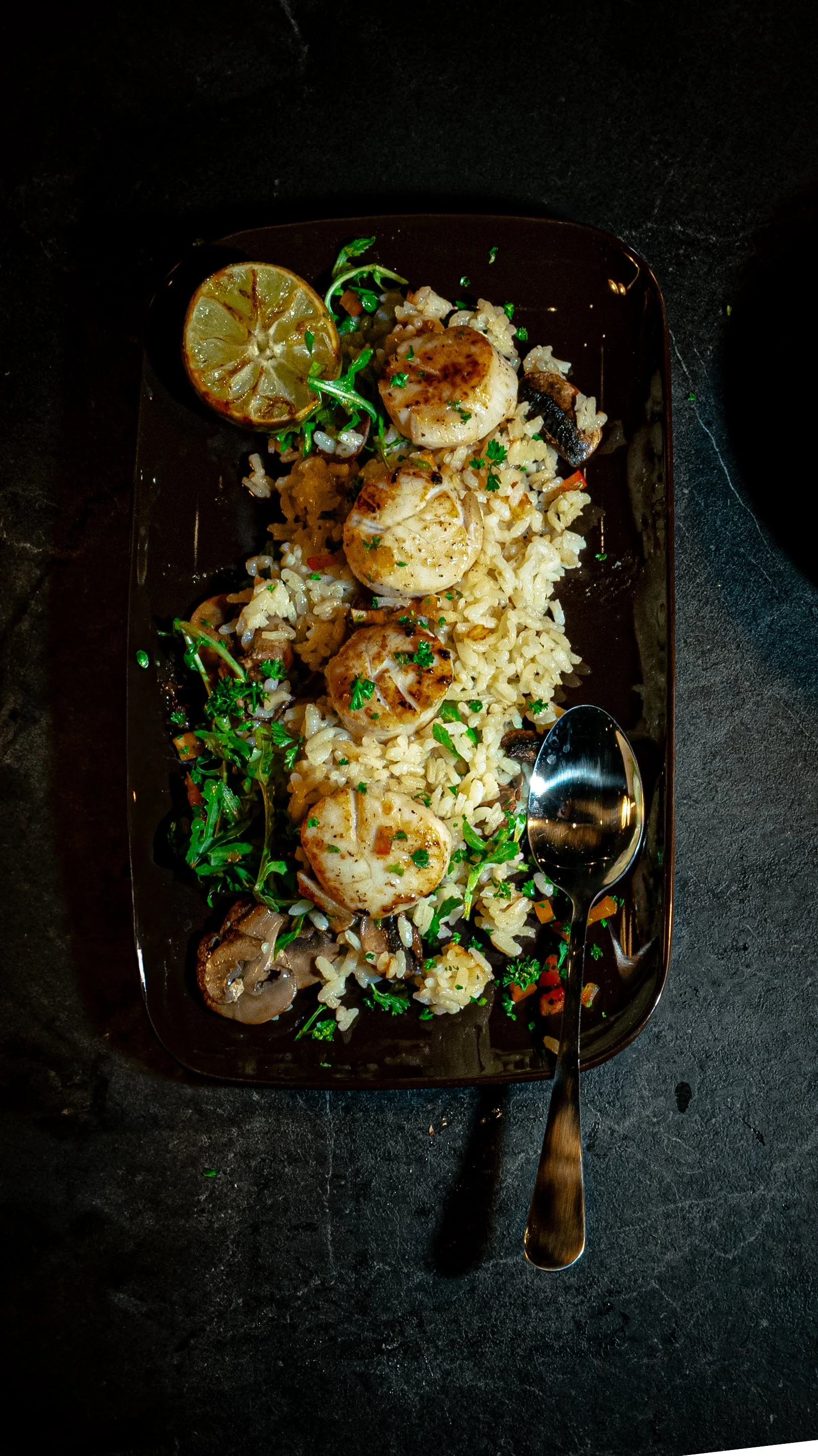 A black rectangular plate with grilled scallops on rice, garnished with parsley, alongside a slice of lemon and sautéed mushrooms, and a silver spoon.