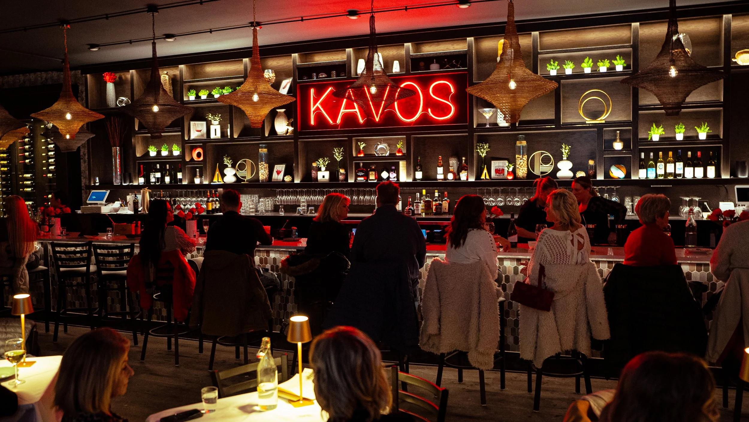 A lively bar with a prominent neon sign reading 'KAVOS' in red, surrounded by patrons sitting at the bar and tables, with dark wood shelves holding bottles, decor, and potted plants, illuminated by warm lighting, including woven pendant lights hangin