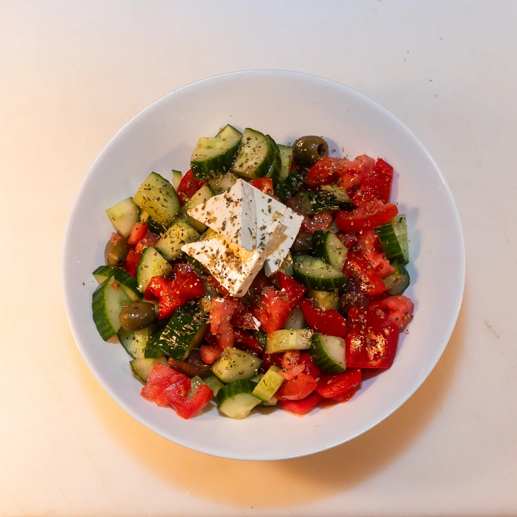 A white bowl filled with a fresh vegetable salad containing chopped cucumbers, tomatoes, red bell peppers, green olives, and a piece of feta cheese, garnished with dried herbs, on a light-colored surface.