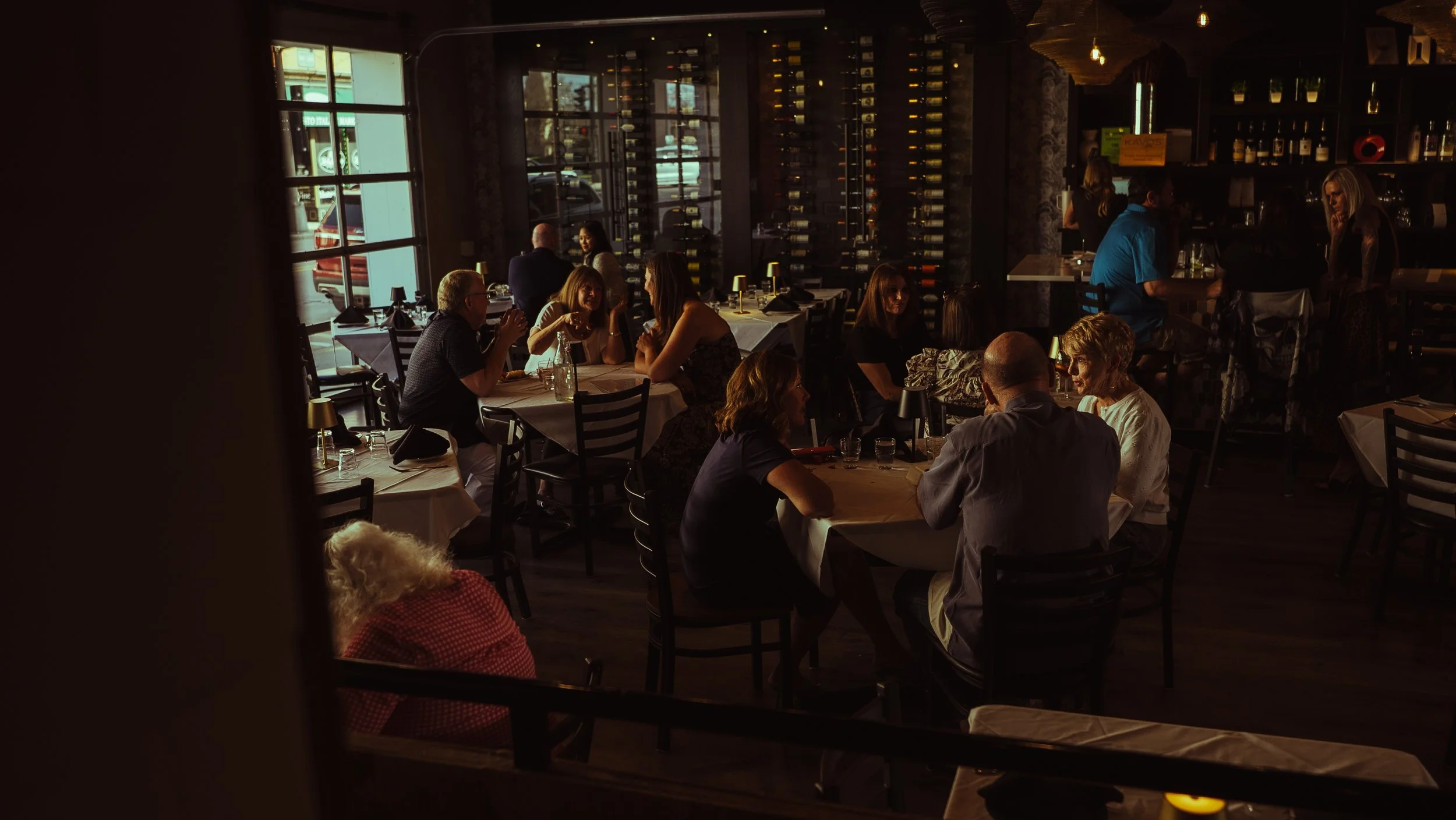 A dimly lit restaurant scene with multiple tables occupied by people dining and conversing. The restaurant features large windows and a wall of wine bottles in the background.