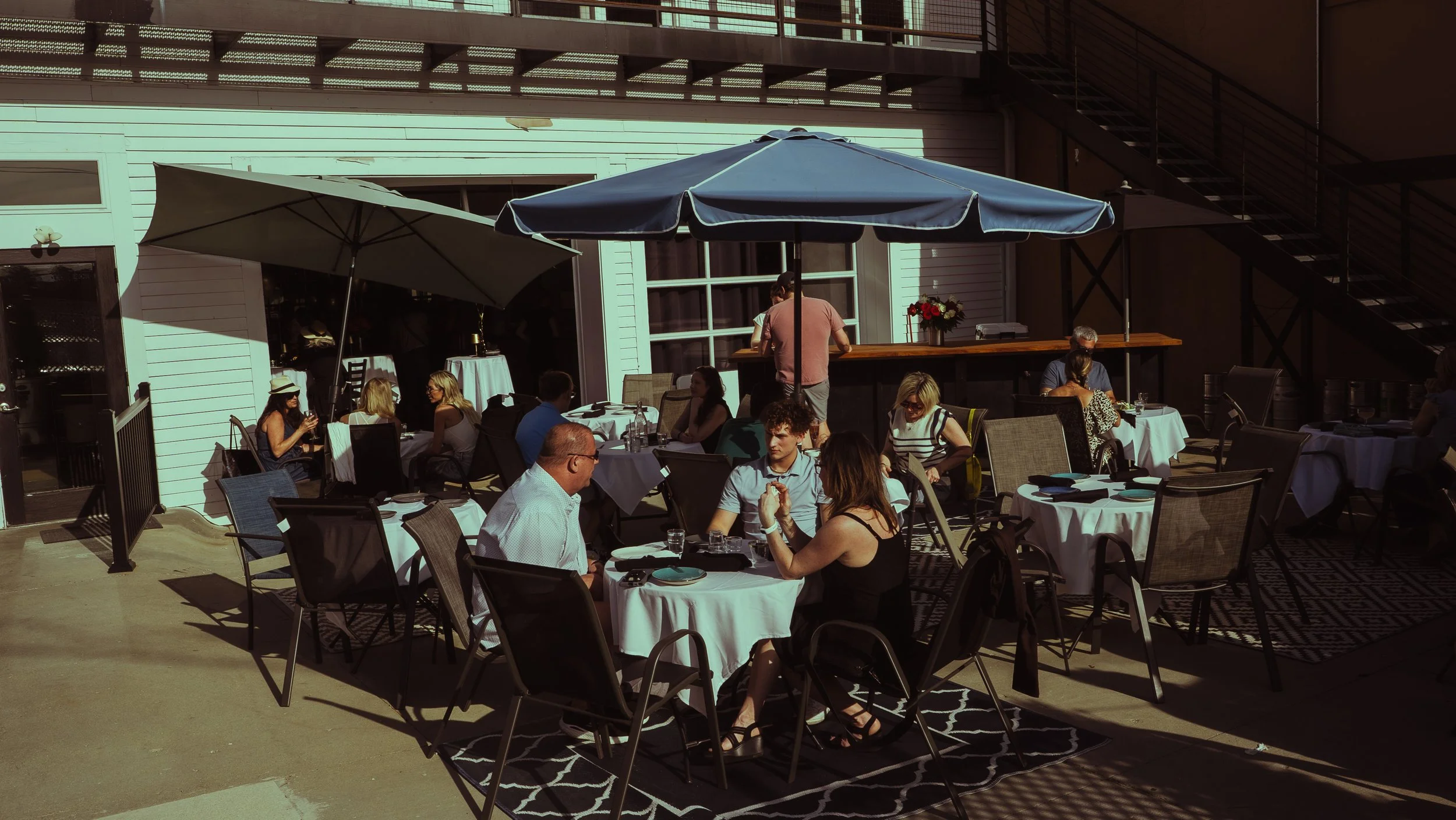 Outdoor rooftop restaurant with people dining at tables with white tablecloths, under large umbrellas, sunny weather, building structure visible in the background.