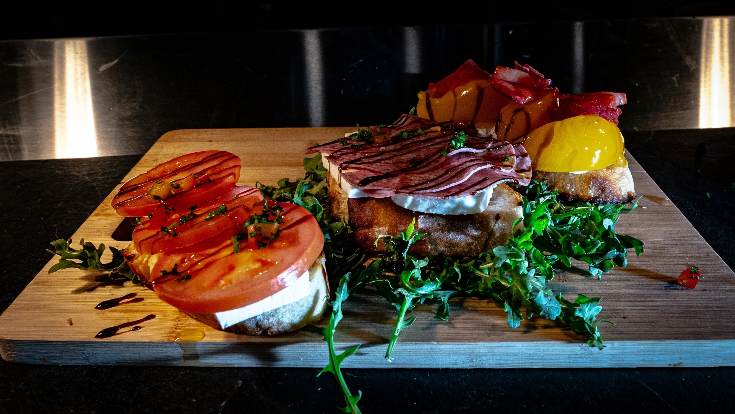 Open-faced sandwiches on a wooden tray garnished with herbs and balsamic glaze, featuring sliced tomatoes, cheese, cured meats, and a yellow pepper, with greens underneath.