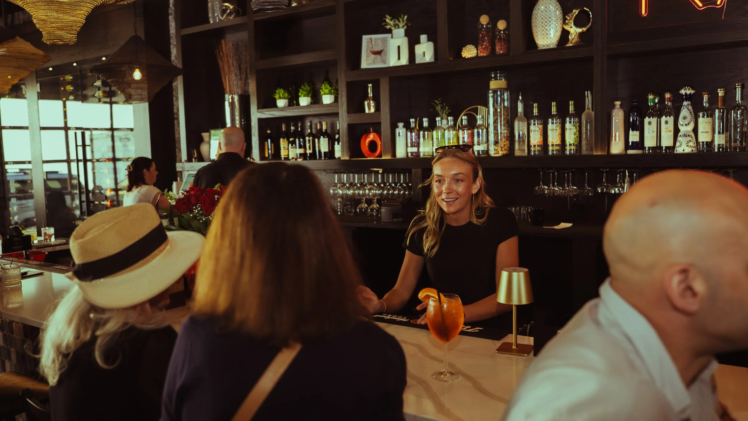 A woman with long blonde hair smiling while talking to a group of people at a bar or restaurant. Various bottles of alcohol and bar decorations are visible in the background, along with a large window letting in natural light.