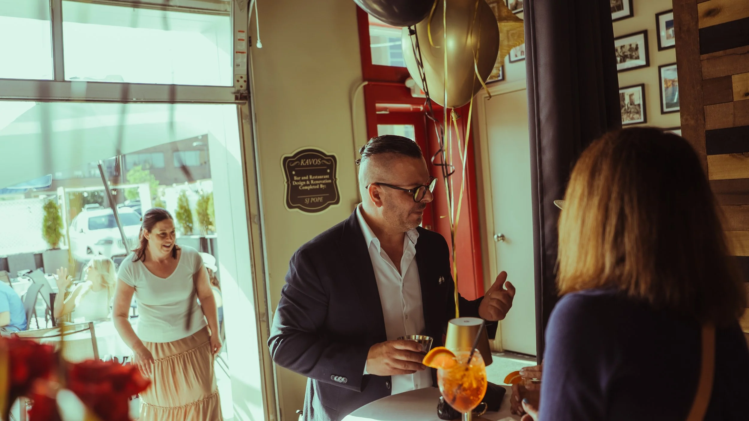 People celebrating inside a restaurant with balloons, including a man in a suit and glasses holding a drink, and a woman with shoulder-length hair.